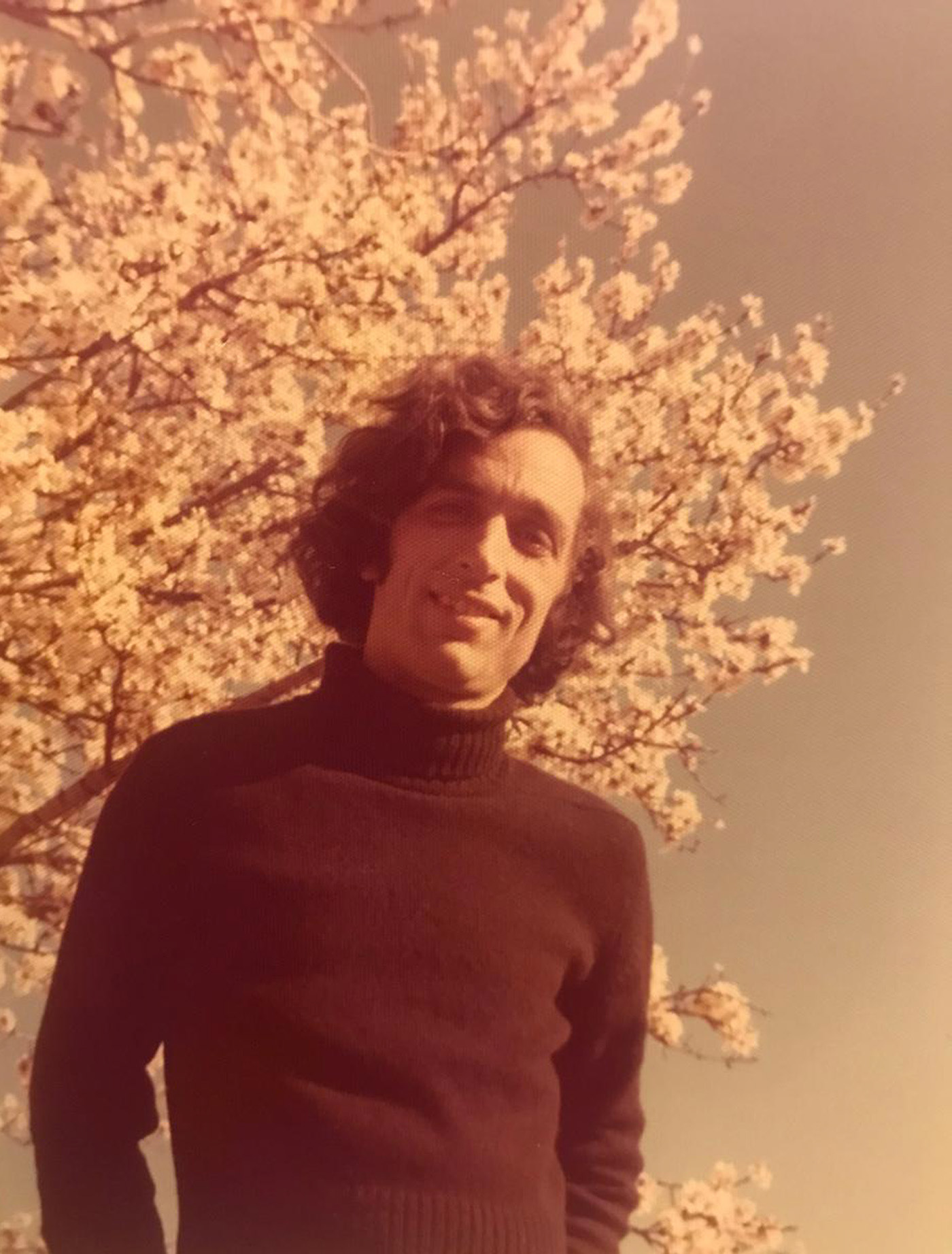 A vintage photograph of a young man standing outdoors, with a backdrop of blossoming trees. The warm, sepia-toned lighting gives the image a nostalgic feel, capturing a moment from the 1970s.