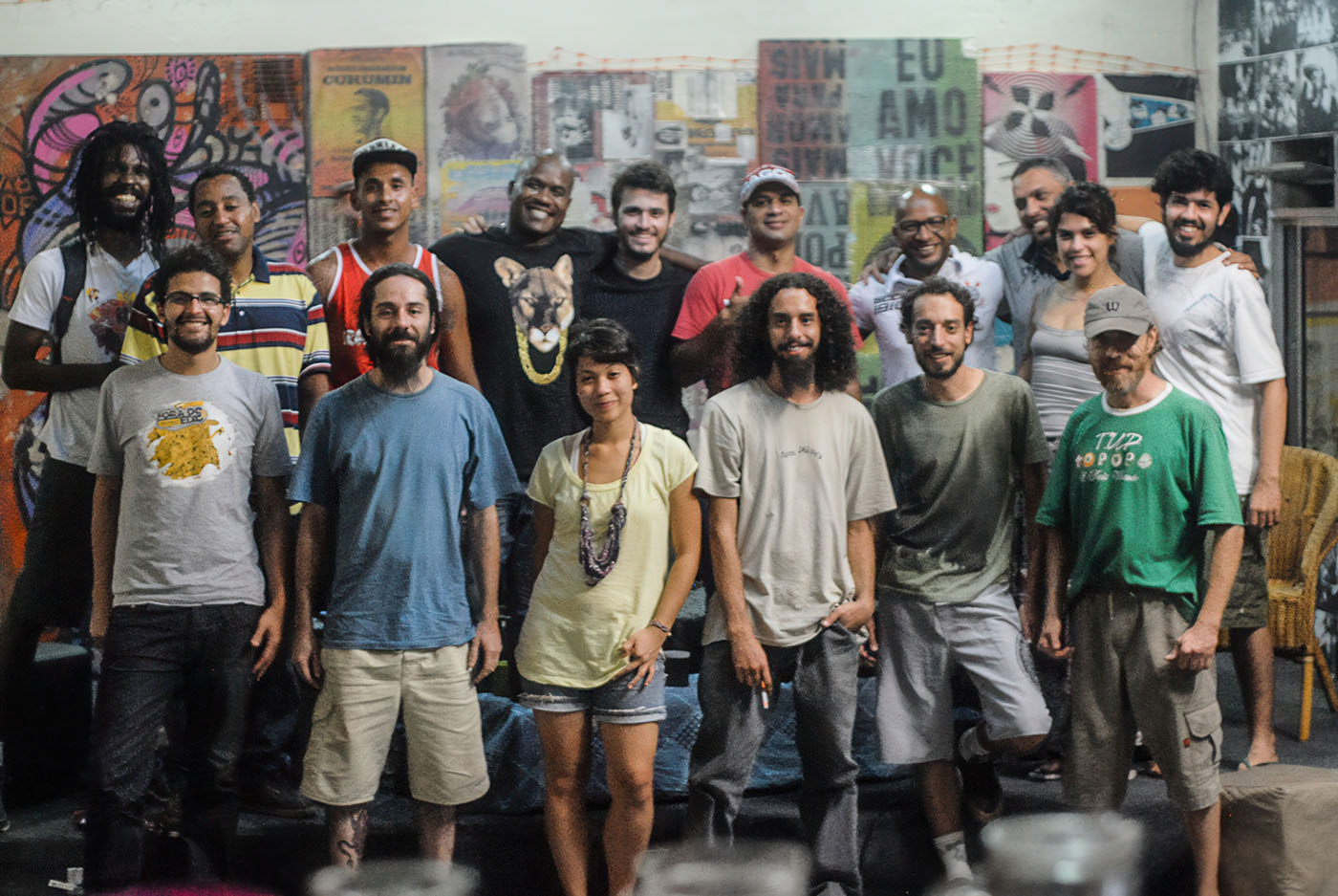 A diverse group of individuals, including men and women, are standing together in a casual pose. They are dressed in various styles of clothing, ranging from t-shirts to shorts, with some wearing hats or accessories. The background features a vibrant mural with bold colors and text, suggesting an artistic or cultural setting. The atmosphere appears relaxed and friendly, indicating a sense of community or shared purpose among the group.