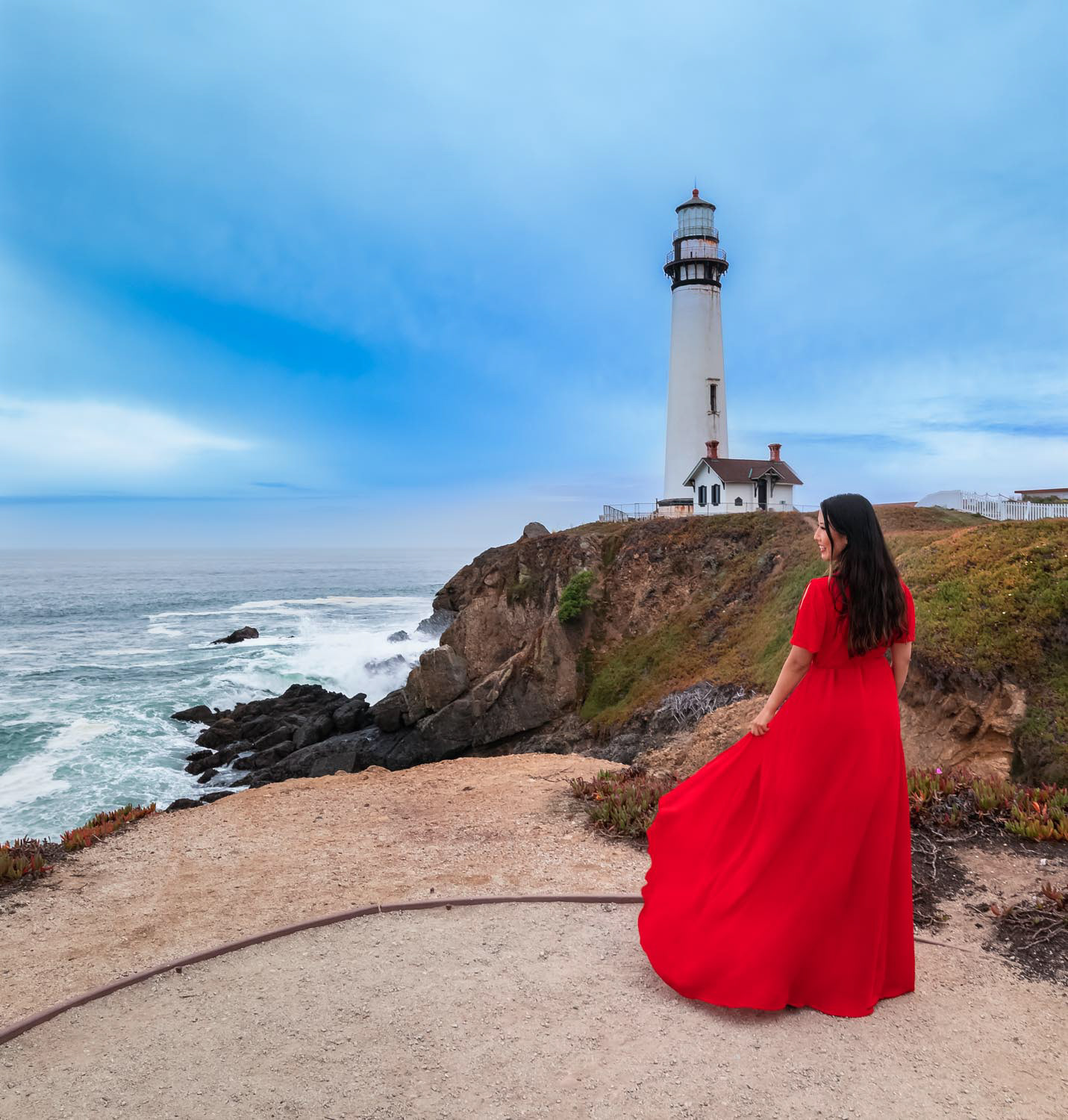 Yong beautiful brunette in a long red dress posing on California Coast with a lighthouse in the background