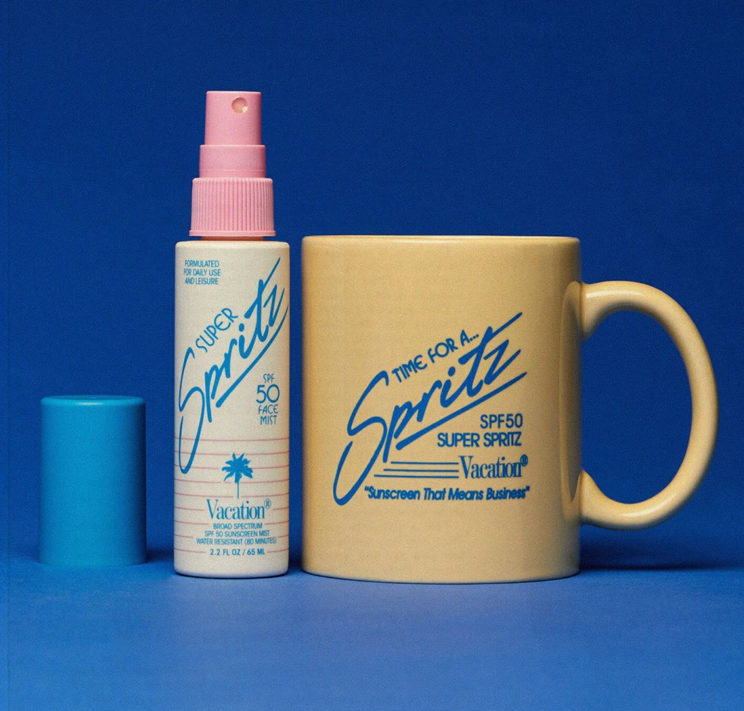 Two of Vacation's products against a bright, dark blue background. It contains their Super Spritz sunscreen spray, a white container with a pink spray top and blue print. Beside the spray is a faded yellow mug with "Time for a Spritz" printed in blue on it.