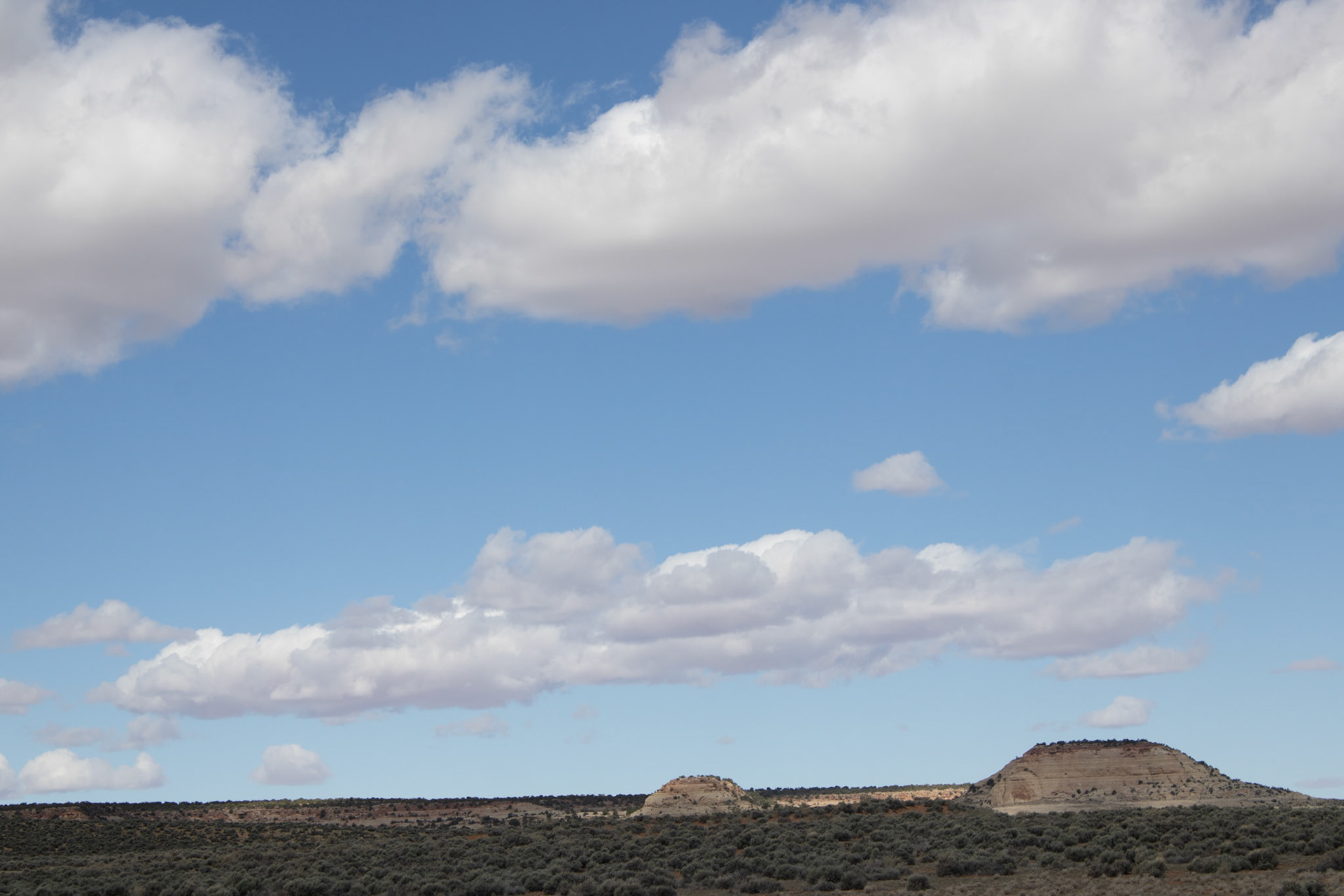 Expansive high desert landscape with layered sandstone hills and sagebrush beneath drifting white cumulus clouds against a clear blue sky in the American West.