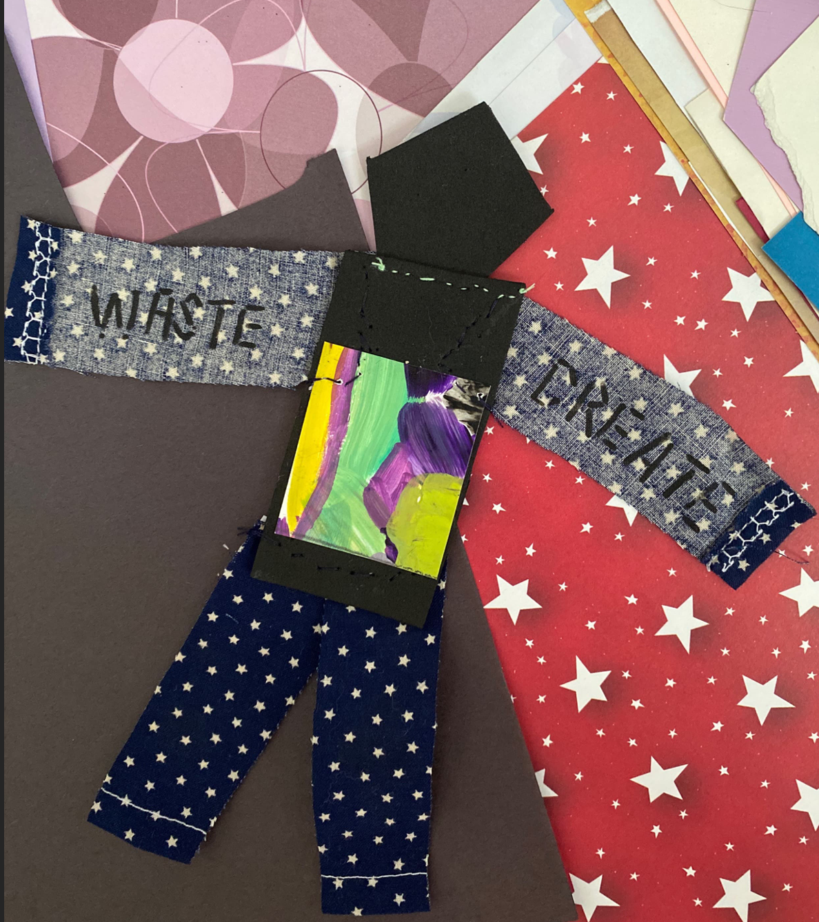 A close-up photo of a paper doll on top of patterned paper. One arm says "Waste" and the other arm says "Create".