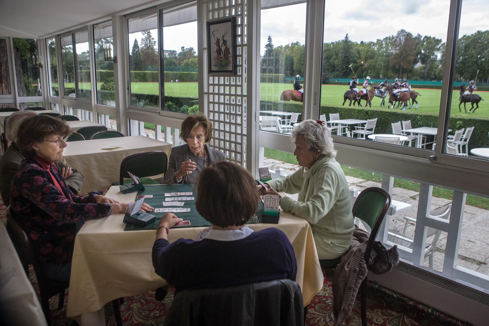 Women, members of the Paris Polo, play bridge as a polo match takes place outside.