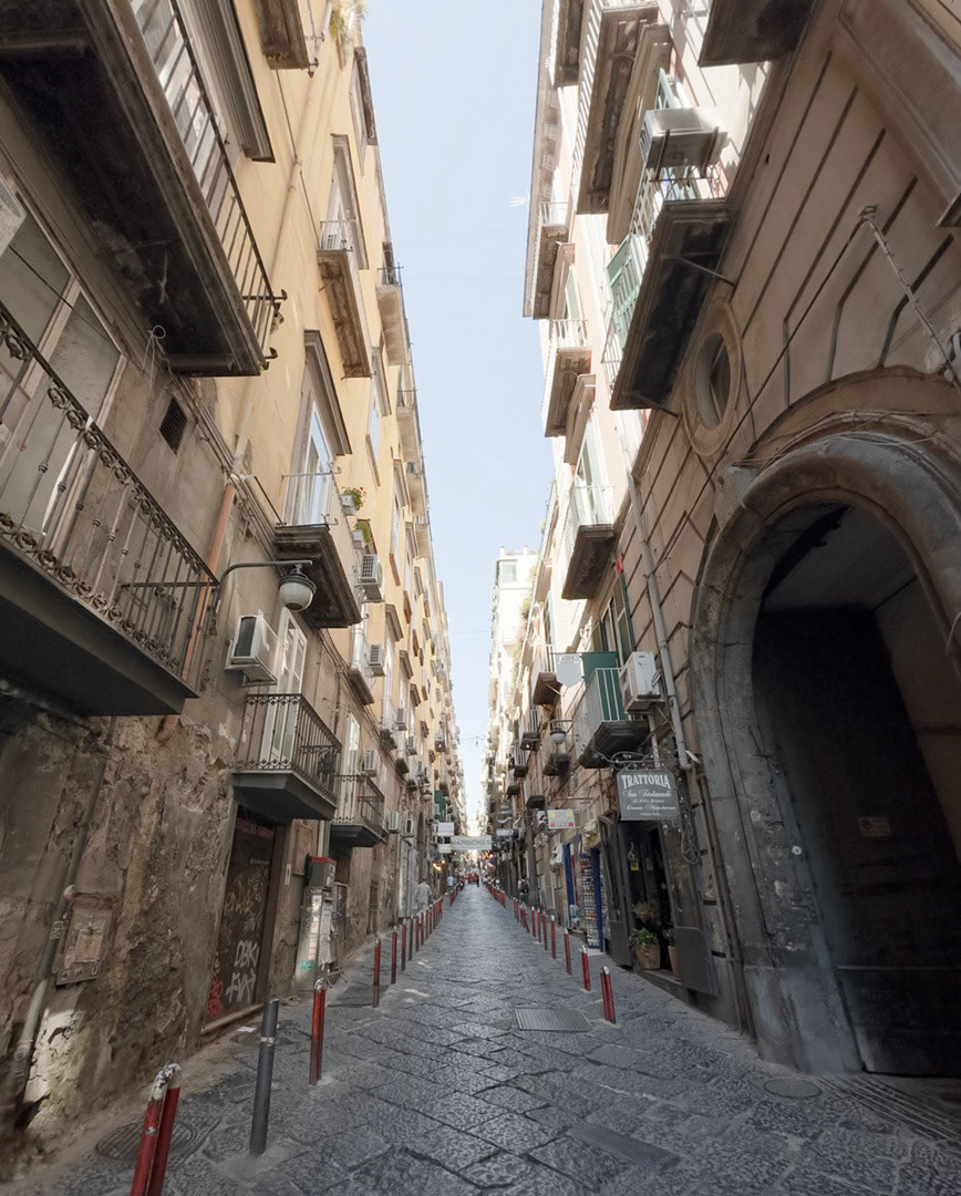 Napoli, Italy - May 27, 2026: Narrow street view along Via Nardones in central Naples, with tall residential buildings, balconies, air conditioners, red bollards, stone paving, shop signs, a trattoria, and strong perspective between weathered historic facades