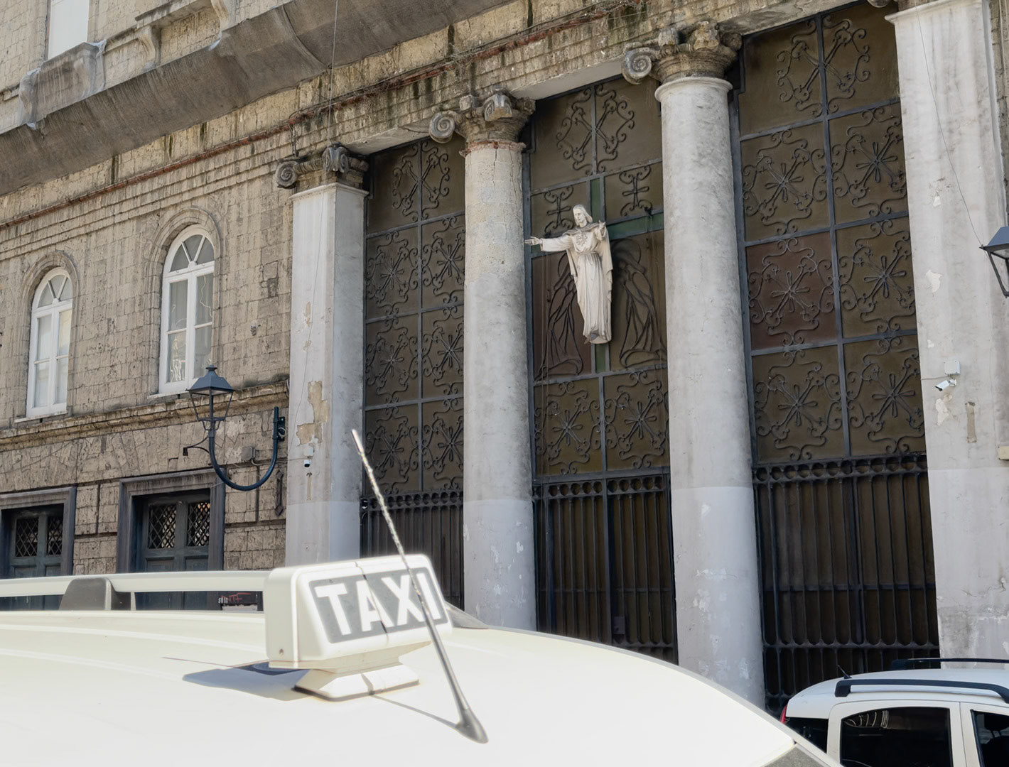 Napoli, Italy - May 27, 2026: Taxi roof sign seen in front of a religious facade on Corso Vittorio Emanuele, with columns, wrought iron gates, a statue of Jesus or the Sacred Heart, and weathered historic architecture