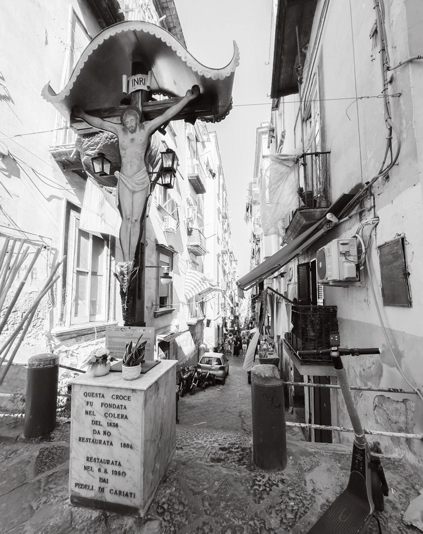 Napoli, Italy - May 27, 2026: Black and white view of a crucifix street shrine in Vico Croce a Cariati, founded during the 1836 cholera epidemic and later restored, standing at the top of a narrow residential alley with scooters, balconies, hanging laundry, cables, and dense urban life