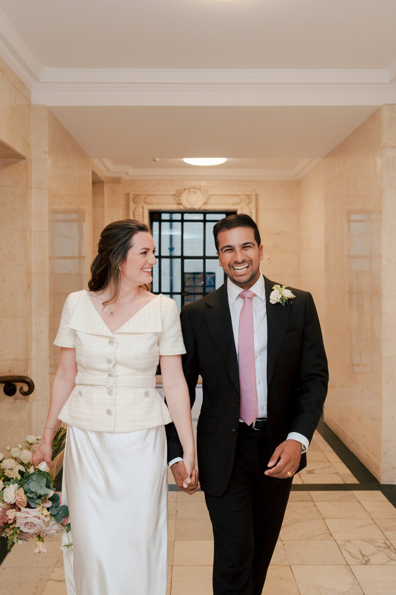 Bride and groom walking hand in hand through the marble hallway of Marylebone Town Hall