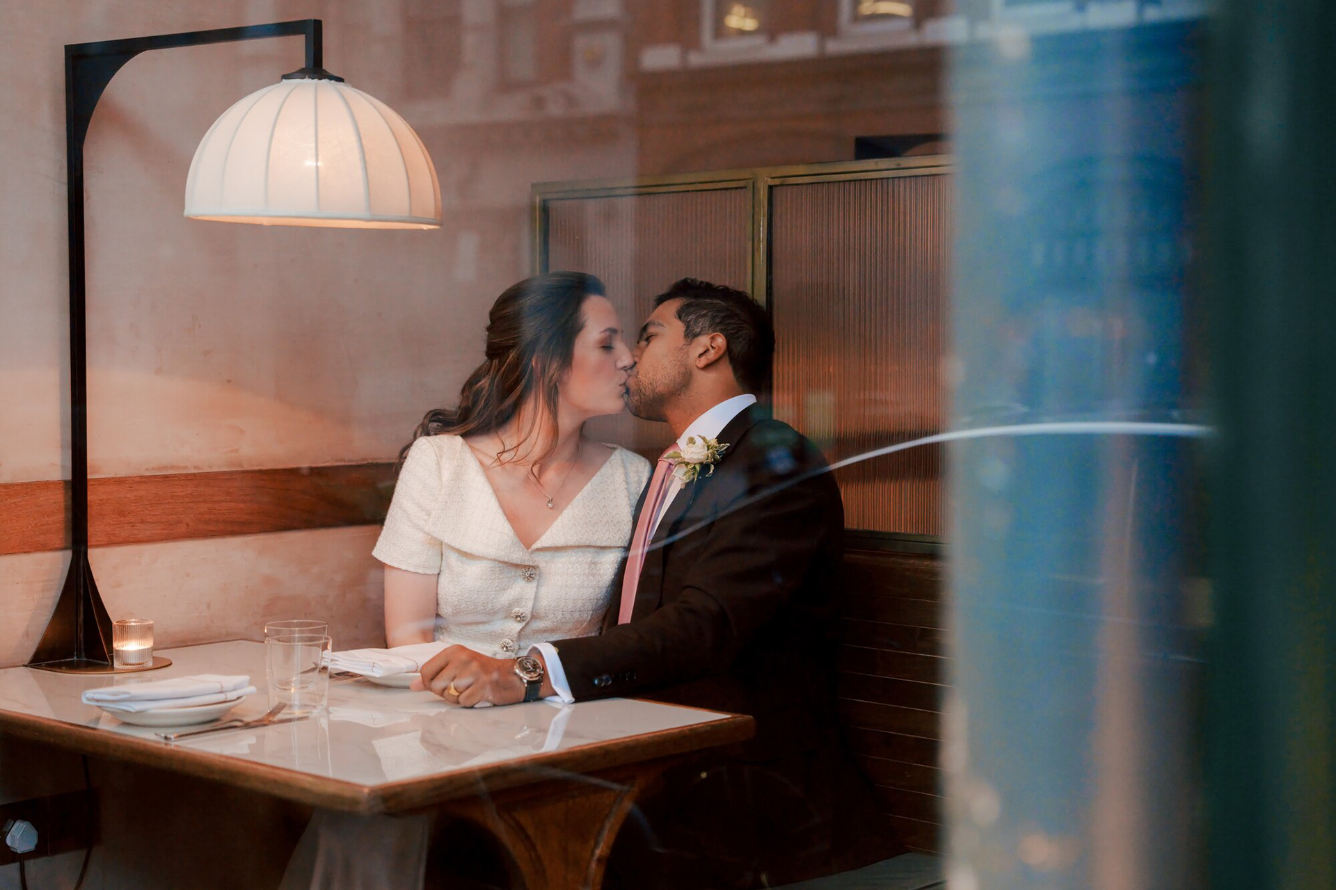 Bride and groom kissing at a table inside Luca restaurant, photographed through the window