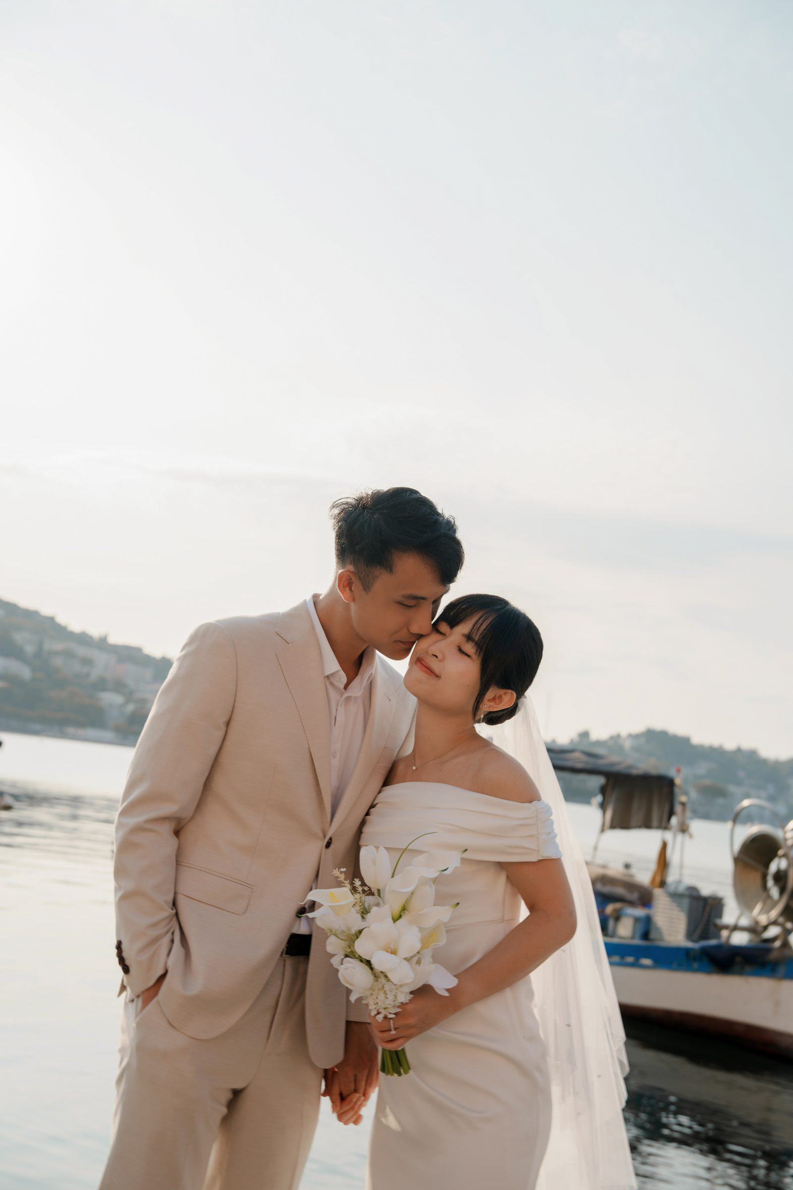 Kiss on the cheek beside the moored boats, bouquet in hand