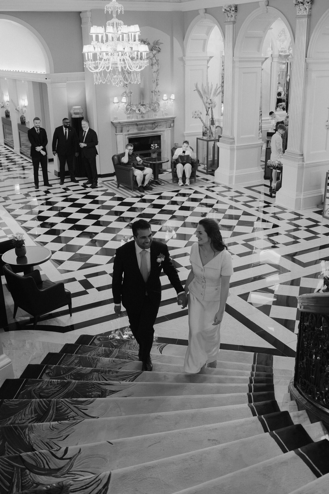 Black and white photo of the couple walking up the chequered marble staircase in the Claridge's lobby