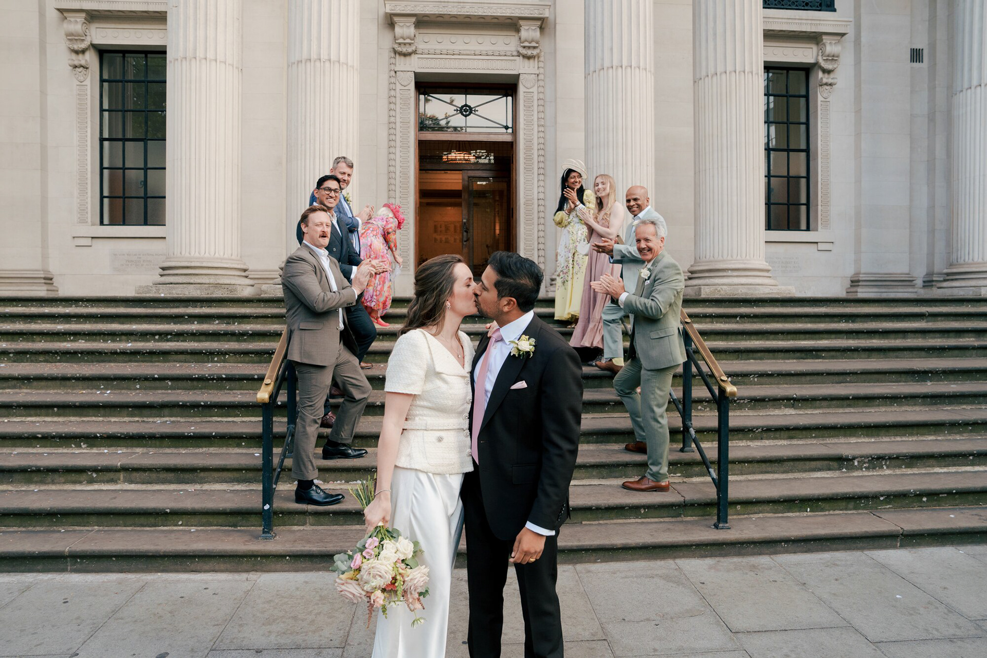 Bride and groom kissing on the steps of Old Marylebone Town Hall as family and friends cheer after their London wedding