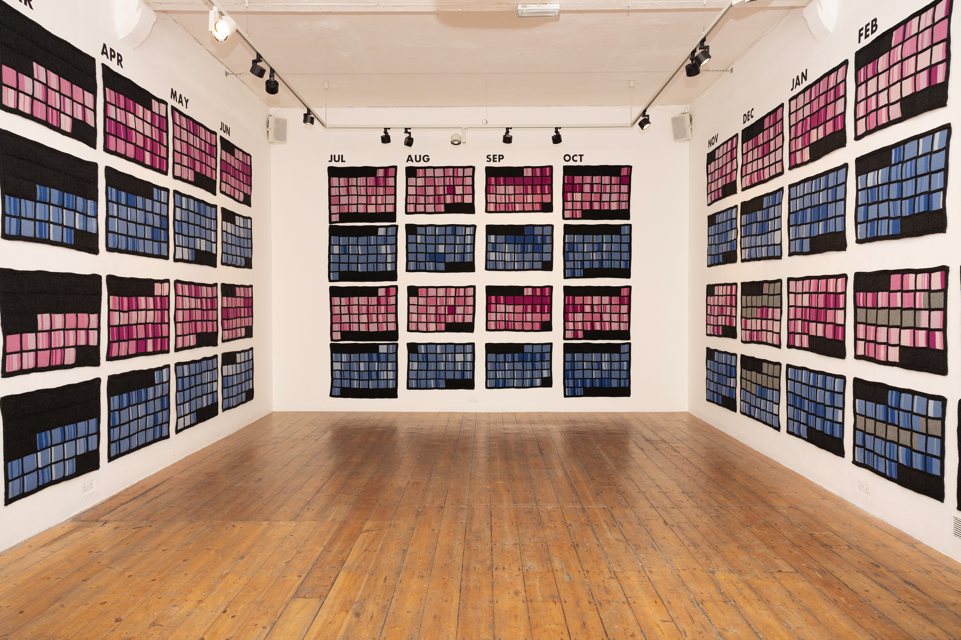 A gallery space featuring a wall installation of colorful textile pieces arranged in a grid format, representing each month with varying shades of pink and blue. The wooden floor complements the artistic display.