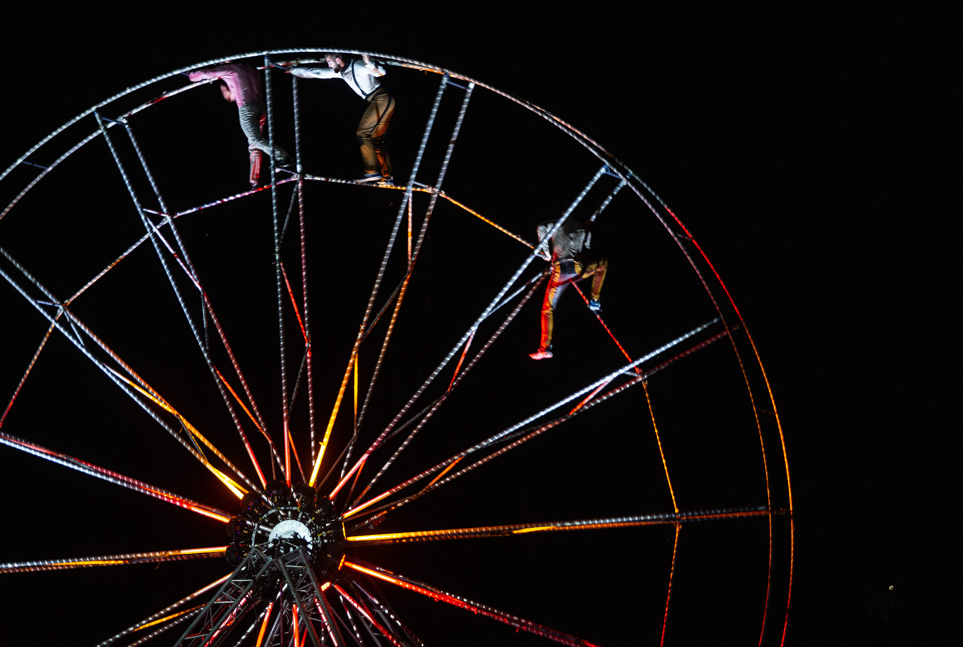 Trois artistes tenant en équilibre à l'intérieur de la grande roue, illuminée dans la nuit.