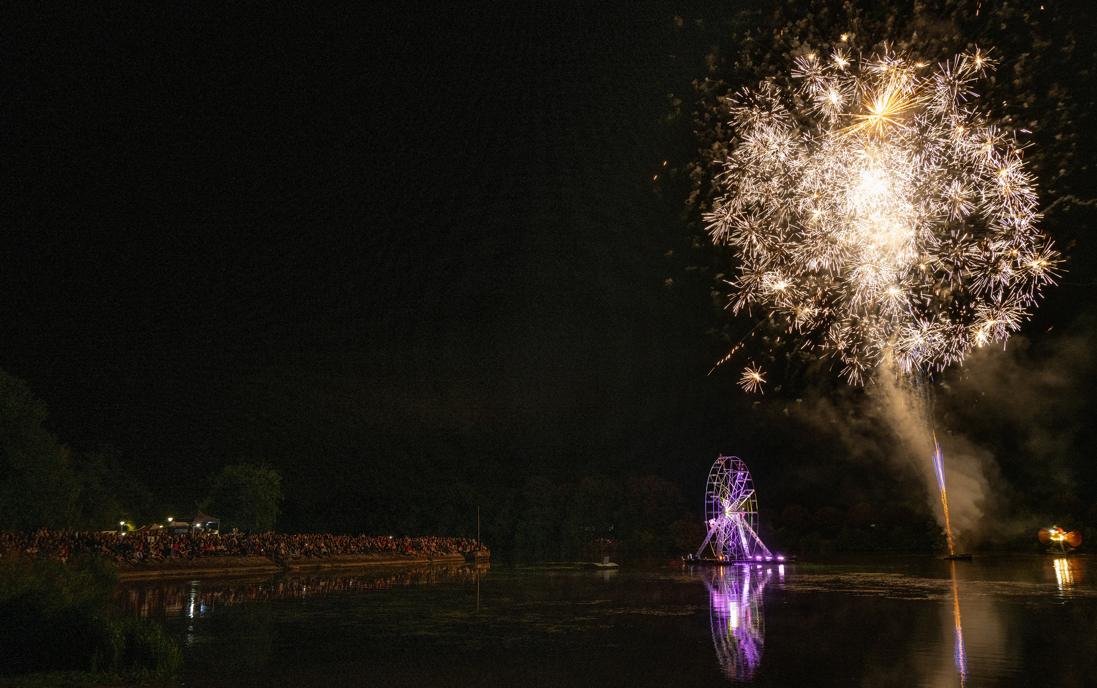 Feux d'artifice éclairant le ciel au-dessus d'une foule rassemblée, avec une grande roue illuminée sur la rive.