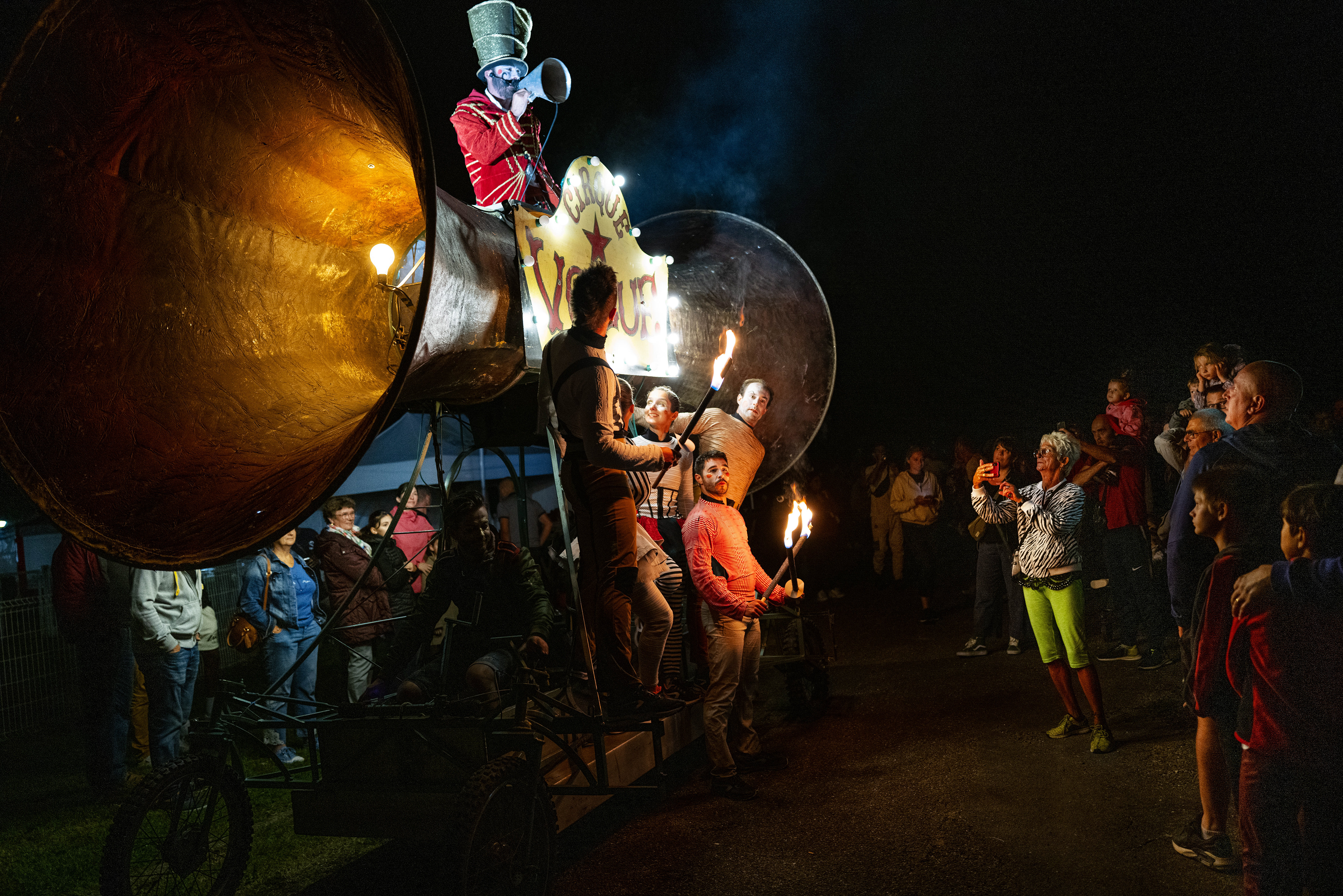 Un spectacle nocturne avec des troubadours autour d'un grand haut-parleur, le maestro en haut, des artistes manipulant des torches de feu, captivant le public.