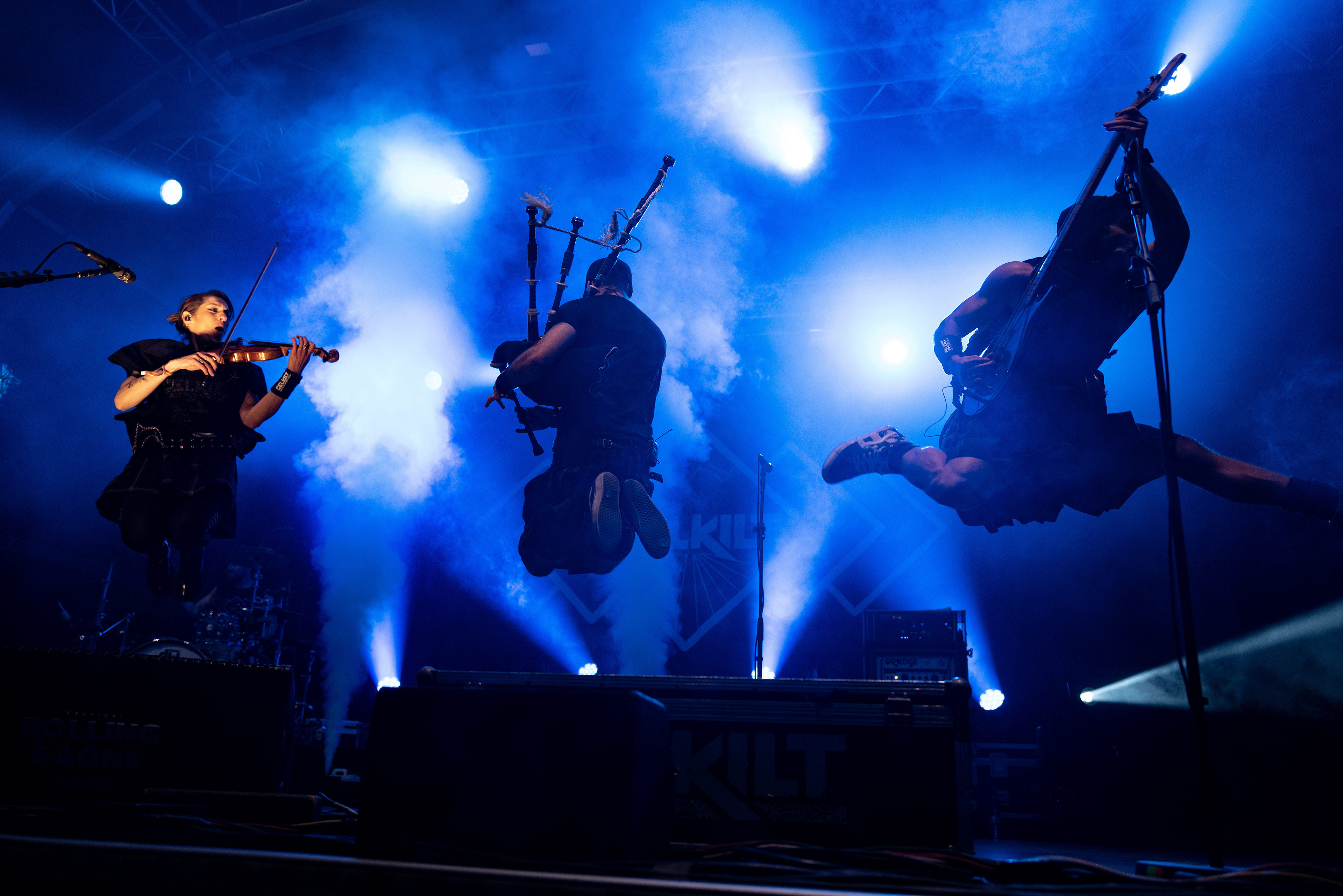 Musiciens du groupe Kilt en train de sauter sur scène, jouant des instruments dans une ambiance de concert illuminée.
