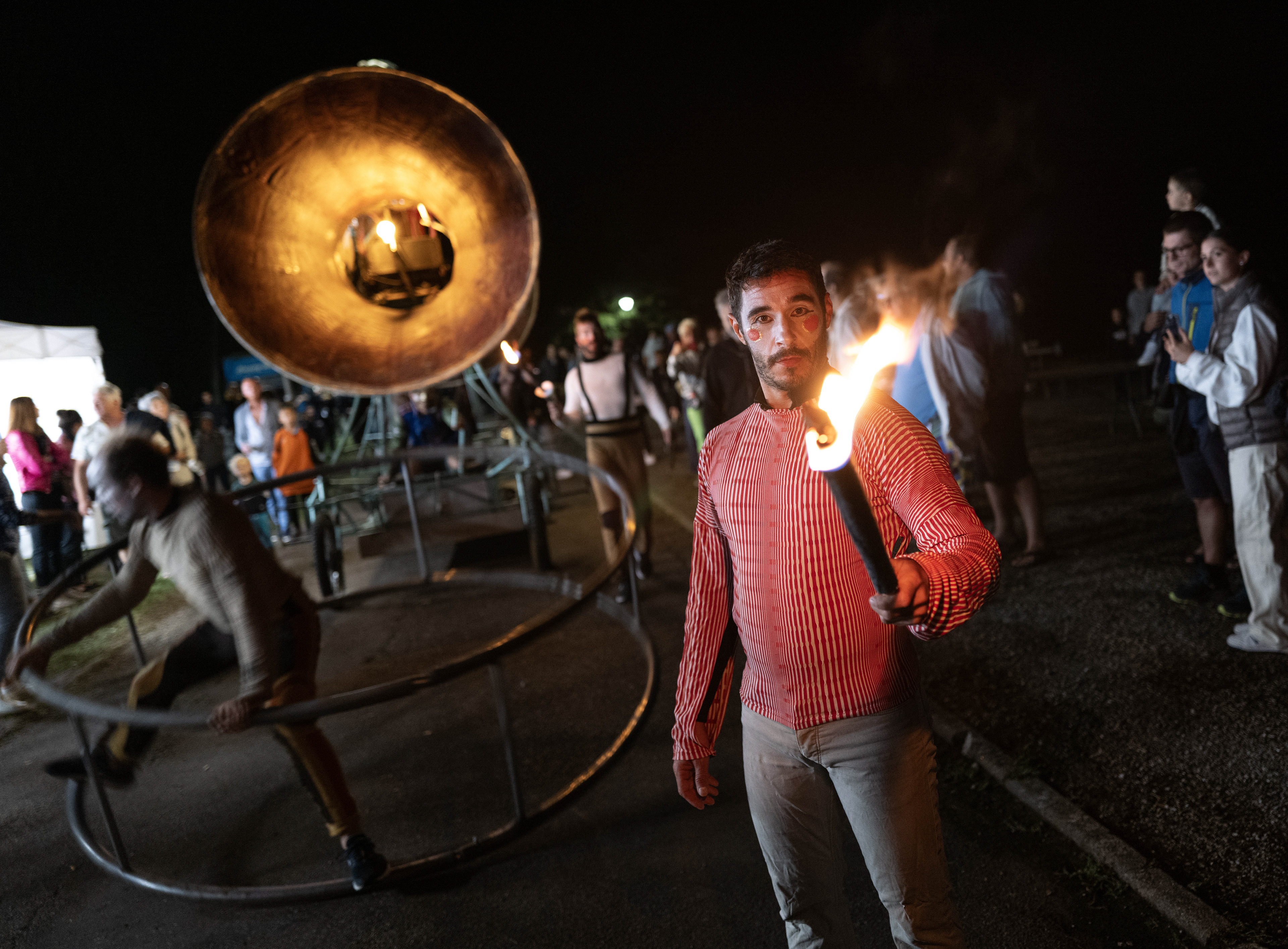 Jeune homme tenant une torche en feu, avec un orchestre de feu en arrière-plan lors d'un spectacle nocturne.