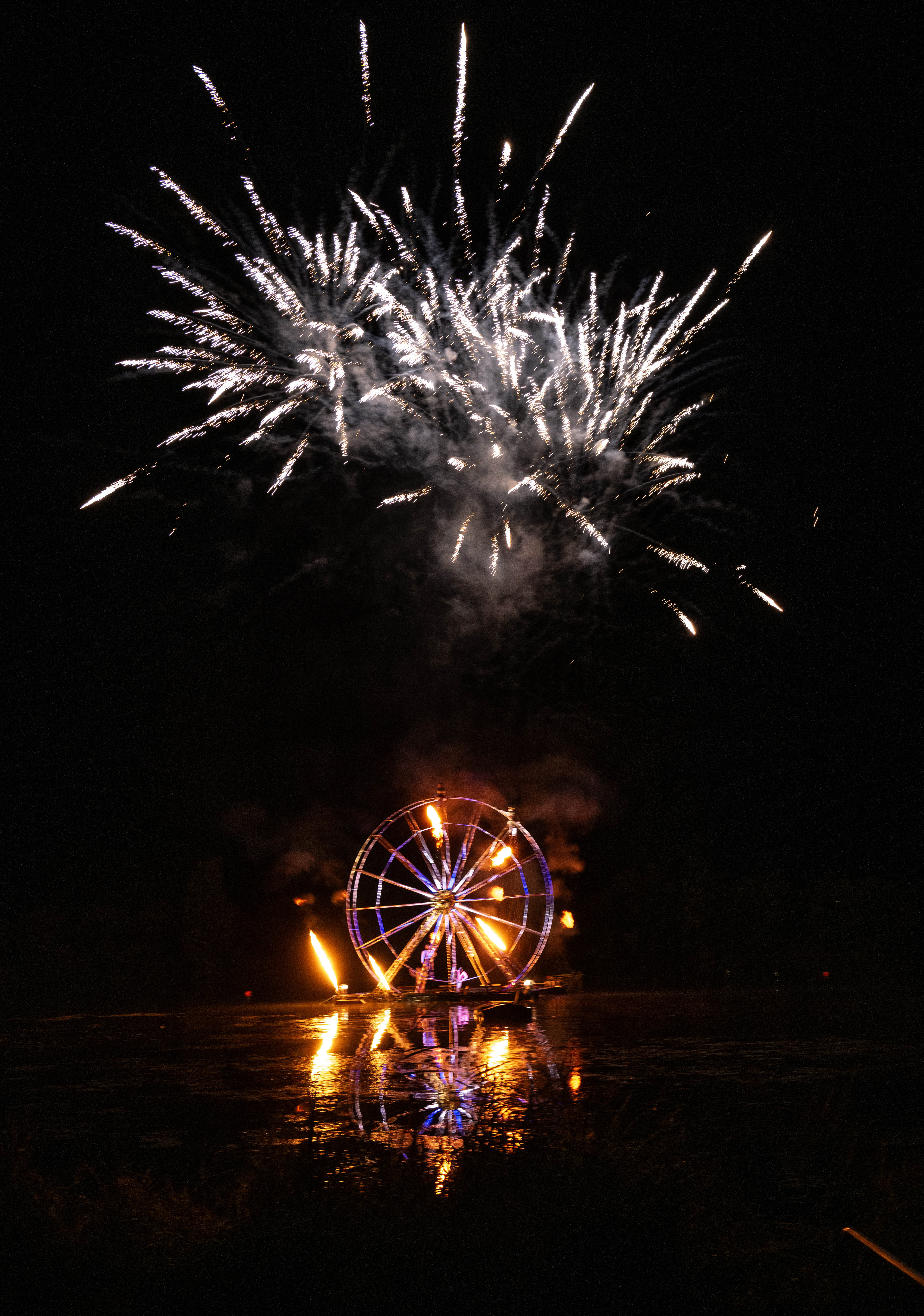 Feux d'artifice éclatant au-dessus d'une grande roue illuminée, avec des reflets scintillants sur l'eau.