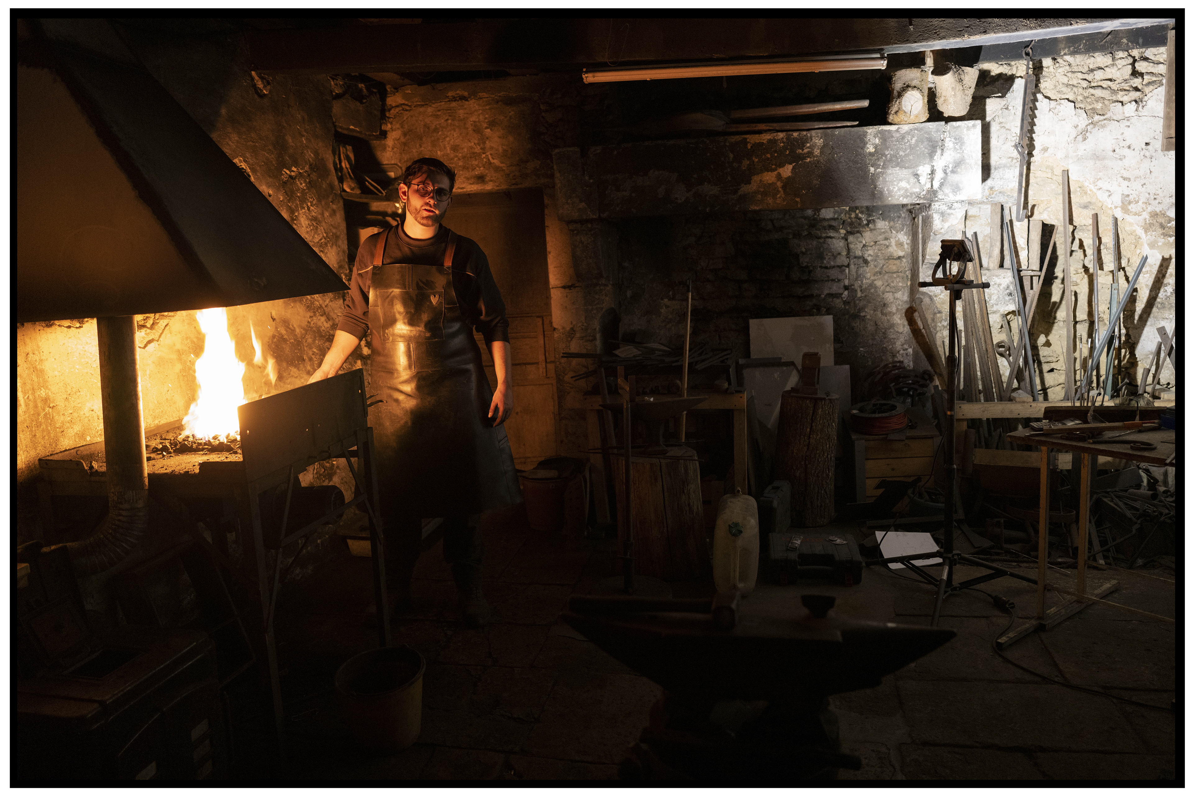 Forgeron dans son atelier ancien, éclairé par un rayon de lumière, entouré de vieux outils de forge.