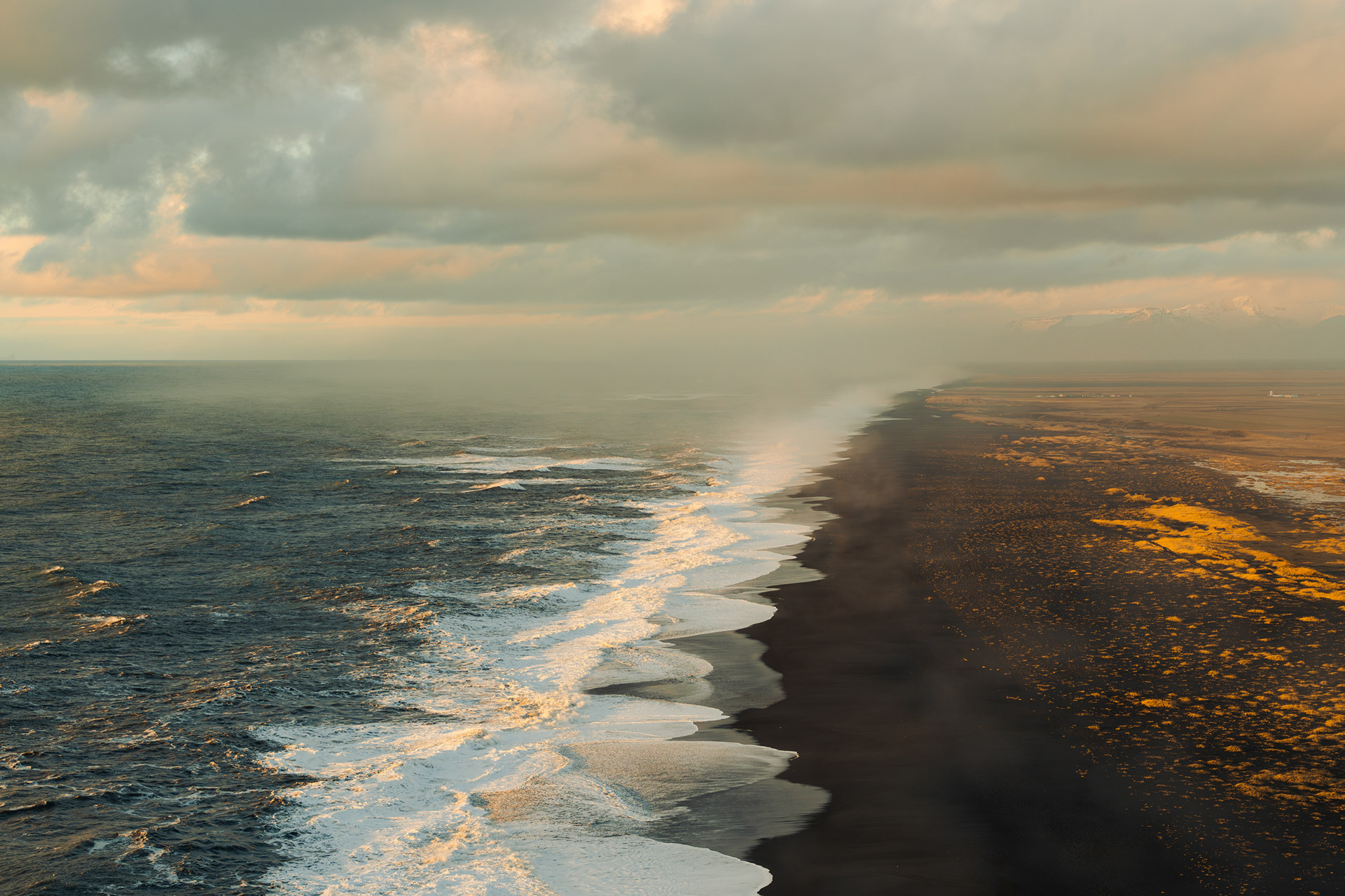 ine art wide shot of Dyrhólaey black sand beach at sunset. The composition is divided into three vertical sections: dark volcanic sand on the right, white ocean foam in the center, and deep blue sea on the left, with subtle orange sunset tones in the cloudy sky highlights