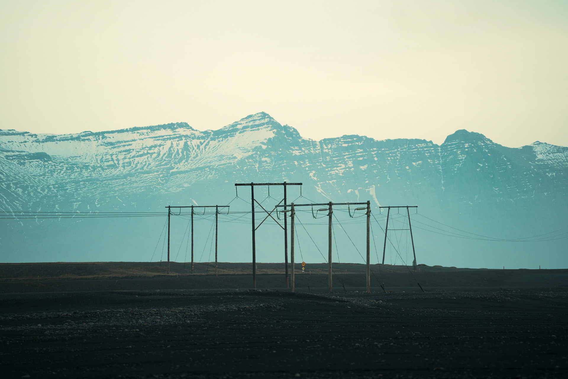 A minimalist, fine art landscape photograph taken near Diamond Beach, Iceland. The image features a series of tall, dark power lines standing starkly against a vast, black volcanic sand plain. In the background, massive blue-toned mountains covered in snow rise under a hazy, pale sky. The composition is clean and cinematic, emphasizing the contrast between human infrastructure and the immense, quiet scale of the Arctic wilderness. Perfectly suited for a professional contact page, symbolizing connection and raw natural beauty.