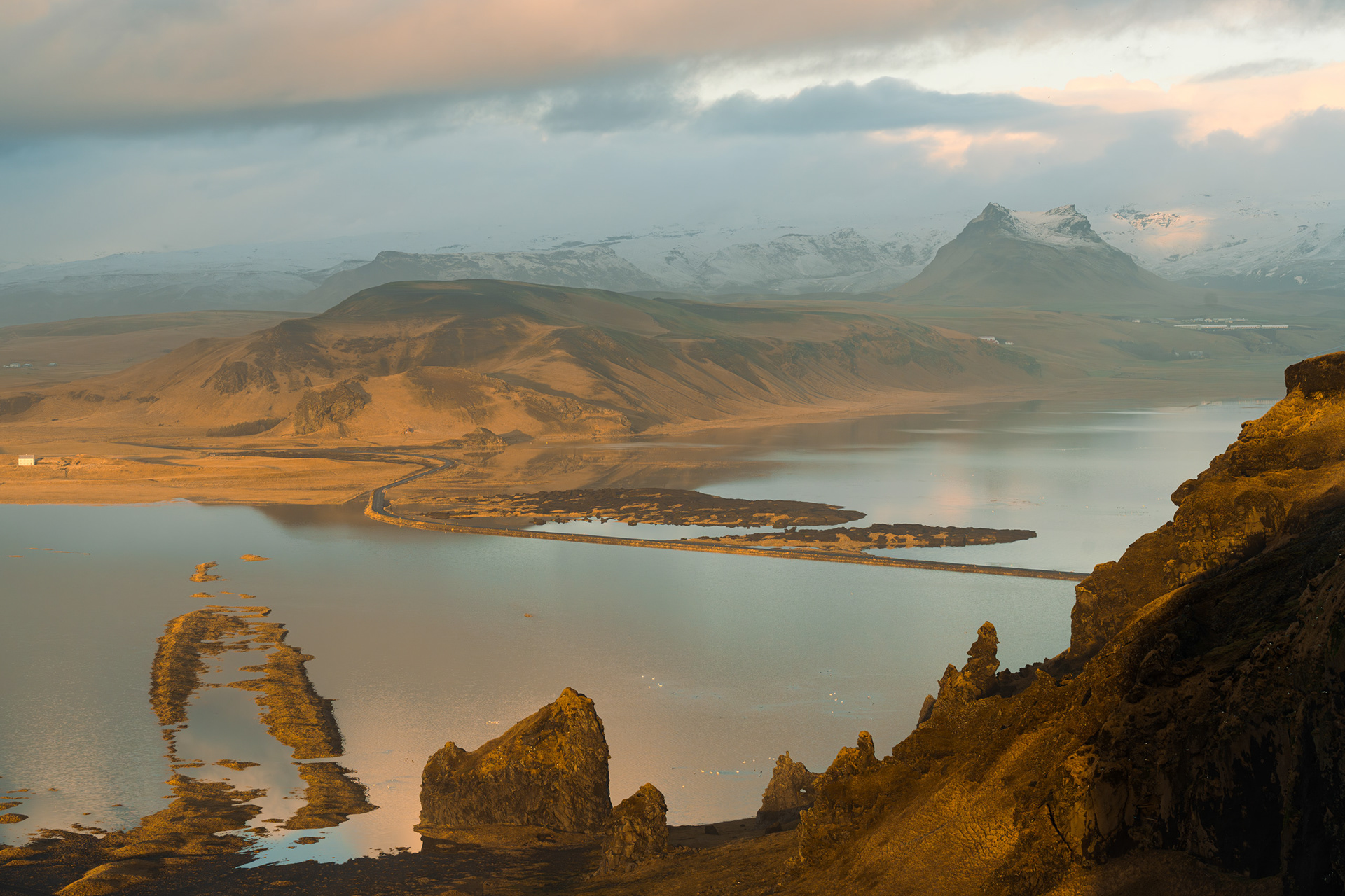 Fine art elevated landscape photograph taken from the cliffs of Dyrhólaey, Iceland. The composition captures the dynamic contrast of golden-hour light illuminating the volcanic sea stacks in the foreground and the snowy, glacier-capped mountains in the distant background. A causeway curves through the shimmering reflection on the black sand lagoon under a cloudy, dramatic sky.