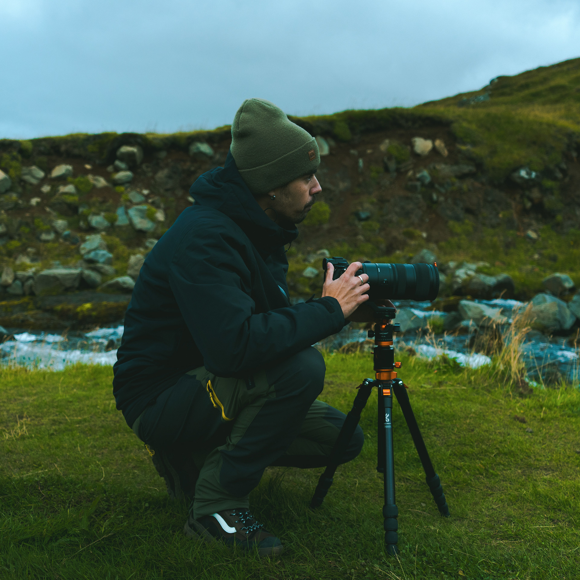 A professional environmental portrait of landscape photographer Daniel Owl(the artist), captured working on location in Iceland. Daniel is crouched on lush green grass, focusing intensely on his camera and telephoto lens mounted on a professional tripod. He is dressed in functional, dark outdoor gear (black hooded jacket, green cargo pants, olive beanie), mirroring the moody colors of the rocky volcanic hill and cloudy sky in the background. A small mountain stream flows nearby. This authentic, atmospheric shot embodies the passion, dedication, and rugged exploration required to capture high-altitude fine art landscapes in remote Arctic environments.