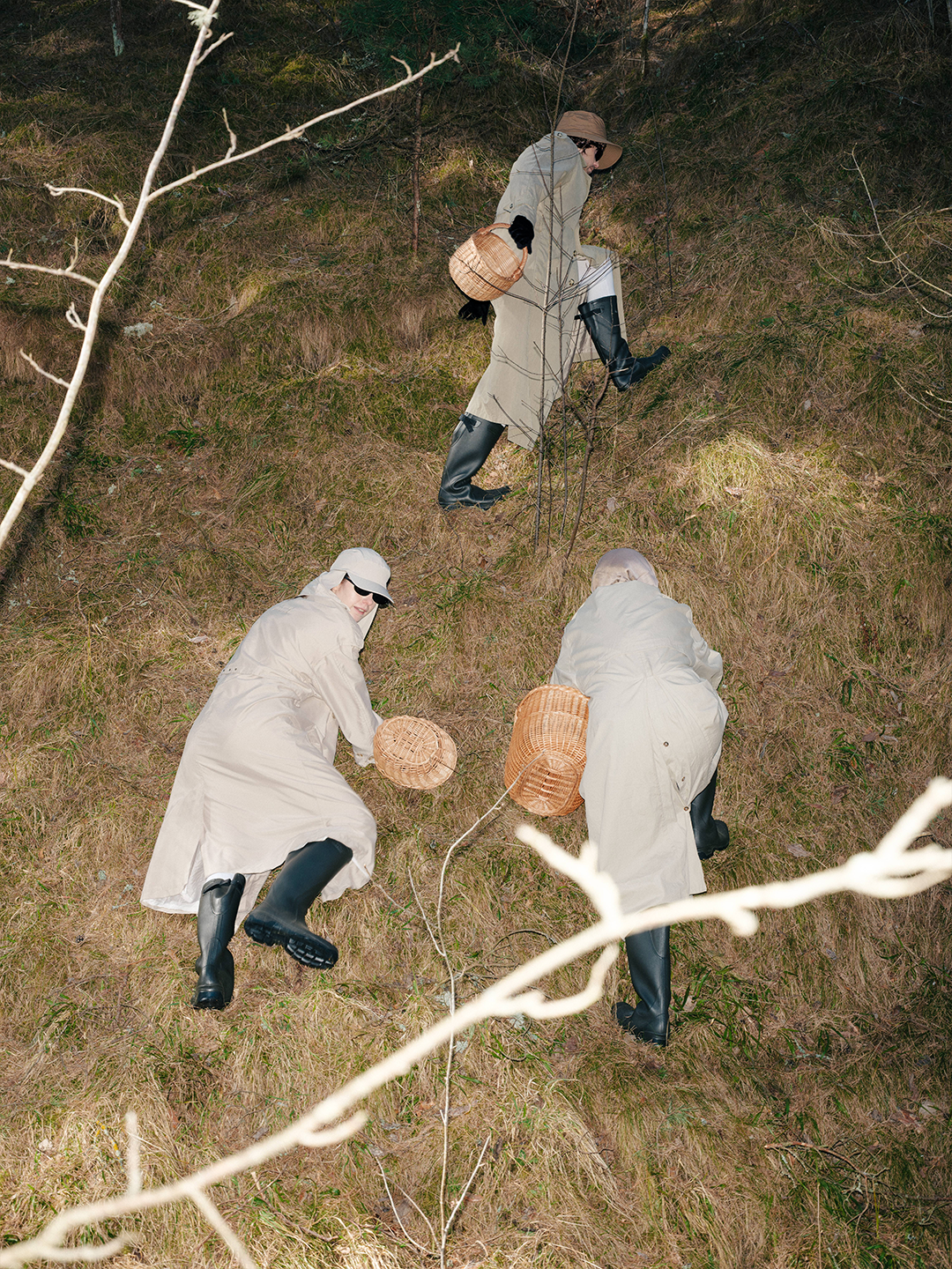 Fine art conceptual photography from a costume design series by Pamela Butāne. High-angle shot of three performers in matching beige trench coats and rain boots foraging with wicker baskets on a grassy hillside.Fine art conceptual photography from a costume design series by Pamela Butāne. High-angle shot of three performers in matching beige trench coats and rain boots foraging with wicker baskets on a grassy hillside.