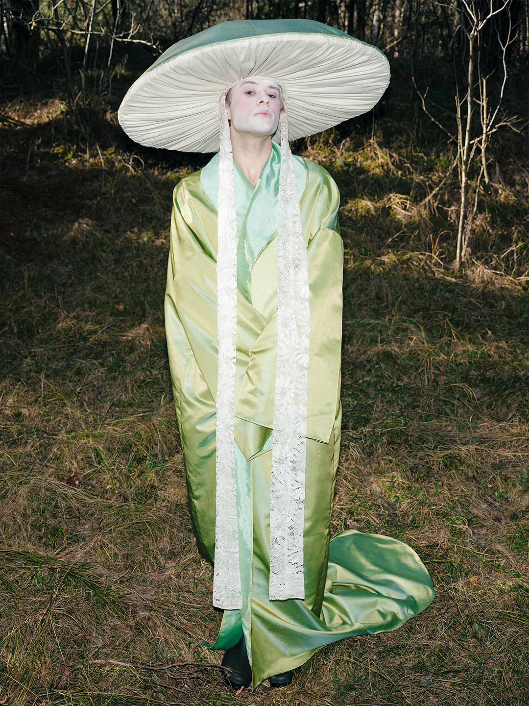 high fashion amanita mushroom costume design by Pamela Butāne. Editorial fashion photography featuring a character in a wide-brimmed green mushroom cap headpiece with lace ties and a draped green satin robe in a forest setting. geisha, aristocratic, rebel