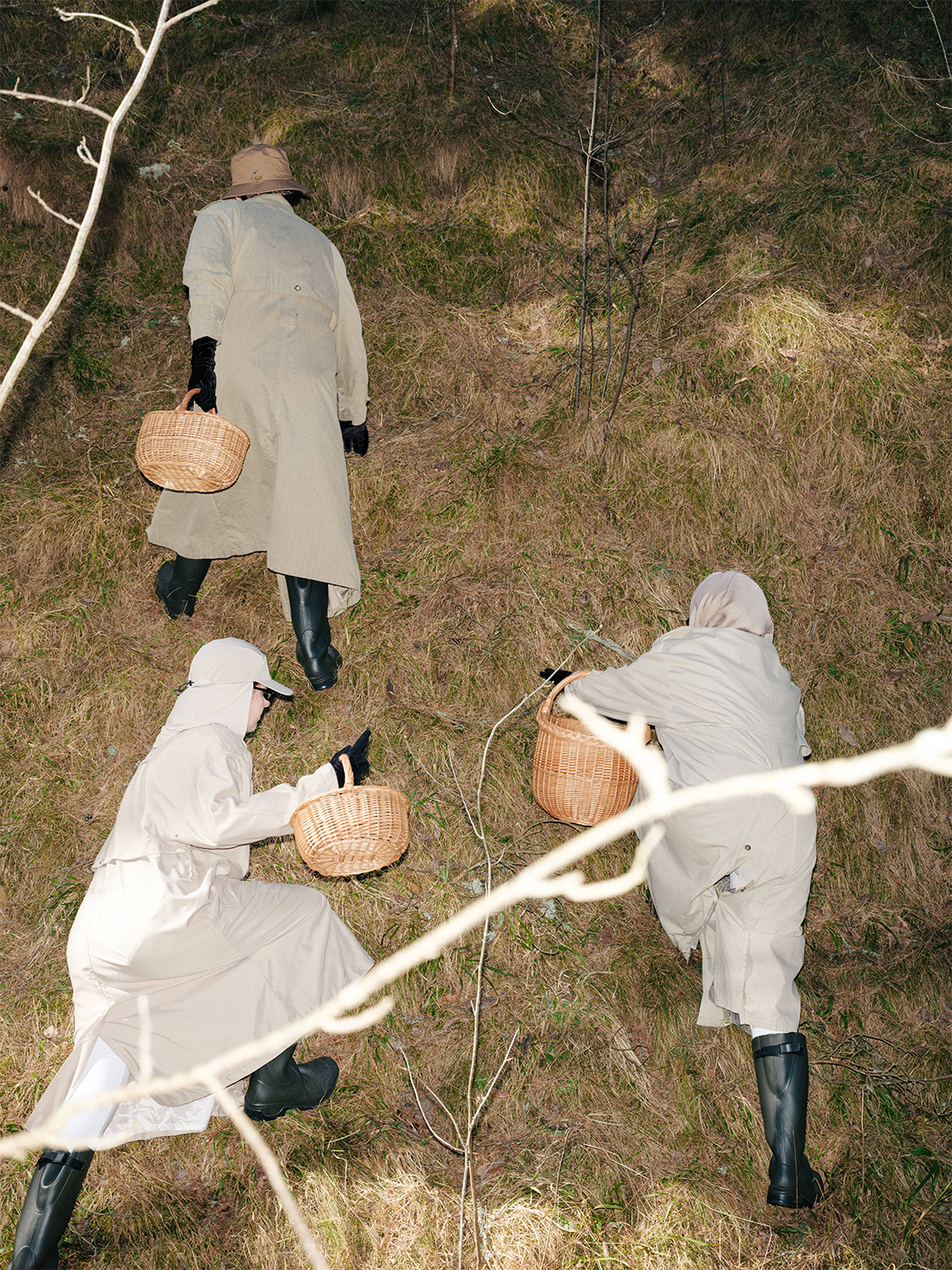 Fine art conceptual photography from a costume design series by Pamela Butāne. High-angle shot of three performers in matching beige trench coats and rain boots foraging with wicker baskets on a grassy hillside. concept author Pamela Butāne- visual artist, scenographer set and costume designer