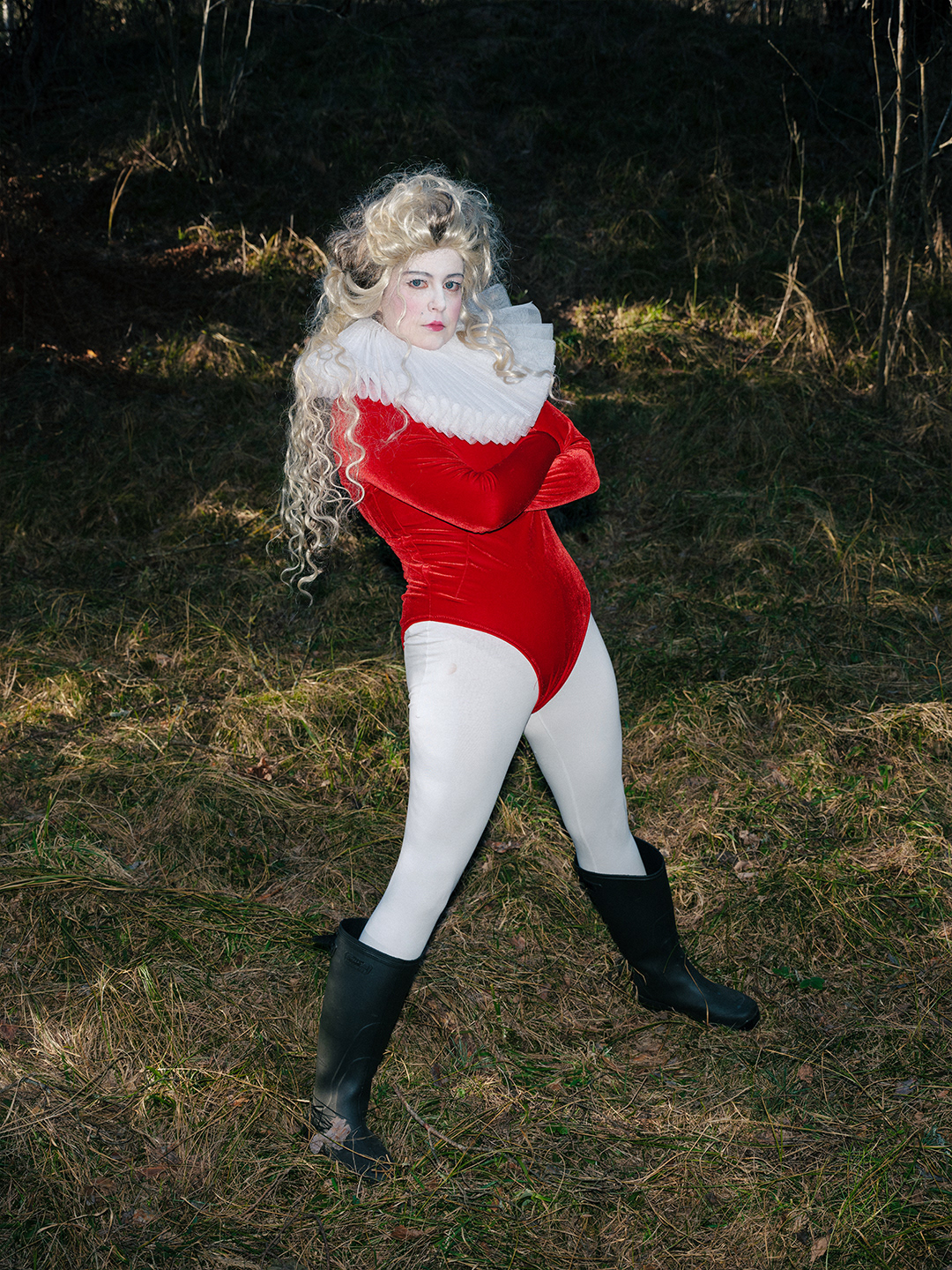 Surreal character costume by designer Pamela Butāne. Studio-style outdoor editorial photography featuring a performer in a white elizabethan ruff collar, a vibrant red velvet bodysuit, and theatrical makeup in the woods. nature and fungi amanita costume by visual artists Pamela Butāne