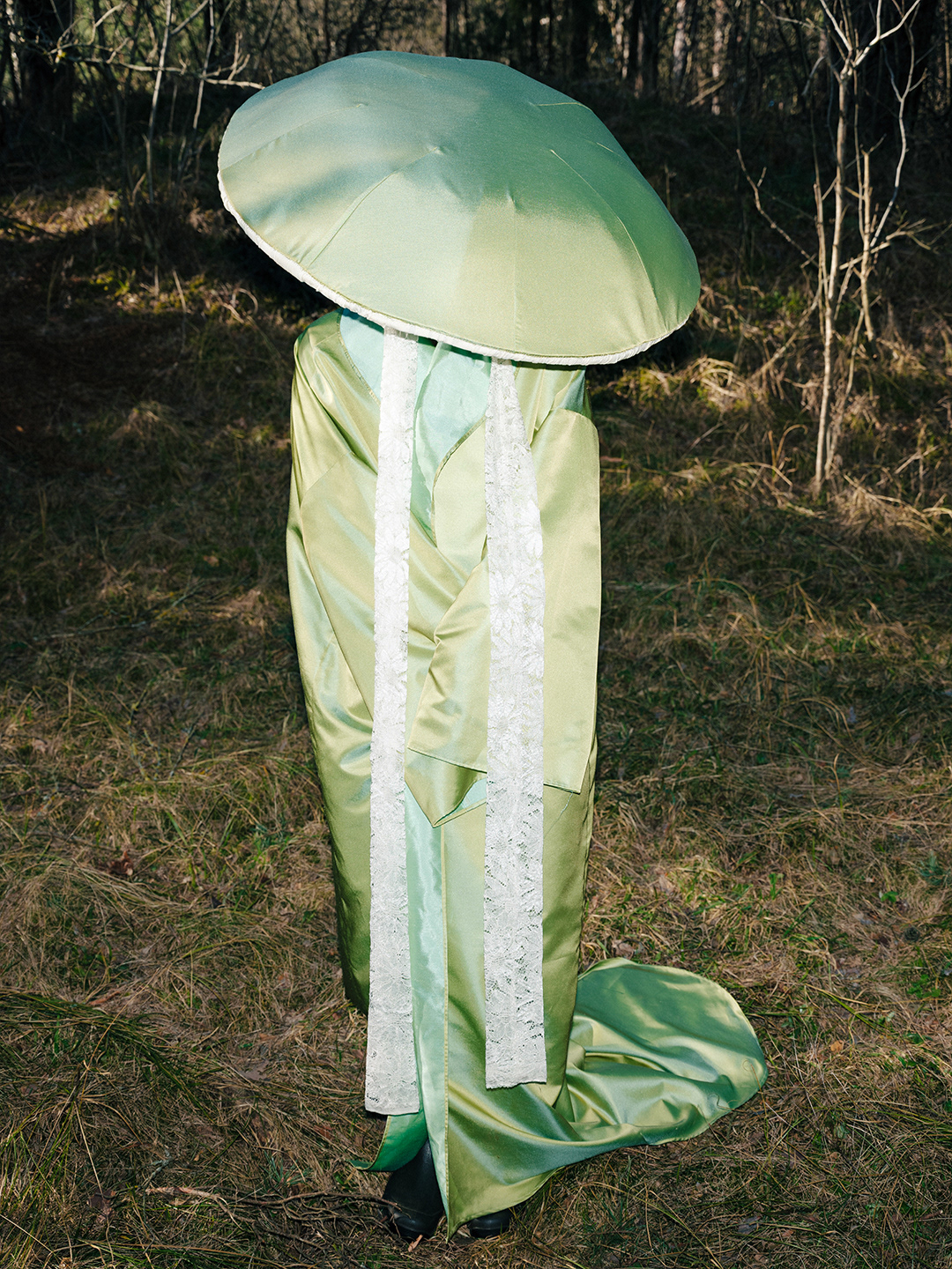 Avant-garde mushroom costume design by Pamela Butāne. Editorial fashion photography featuring a character in a wide-brimmed green mushroom cap headpiece with lace ties and a draped green satin robe in a forest setting.