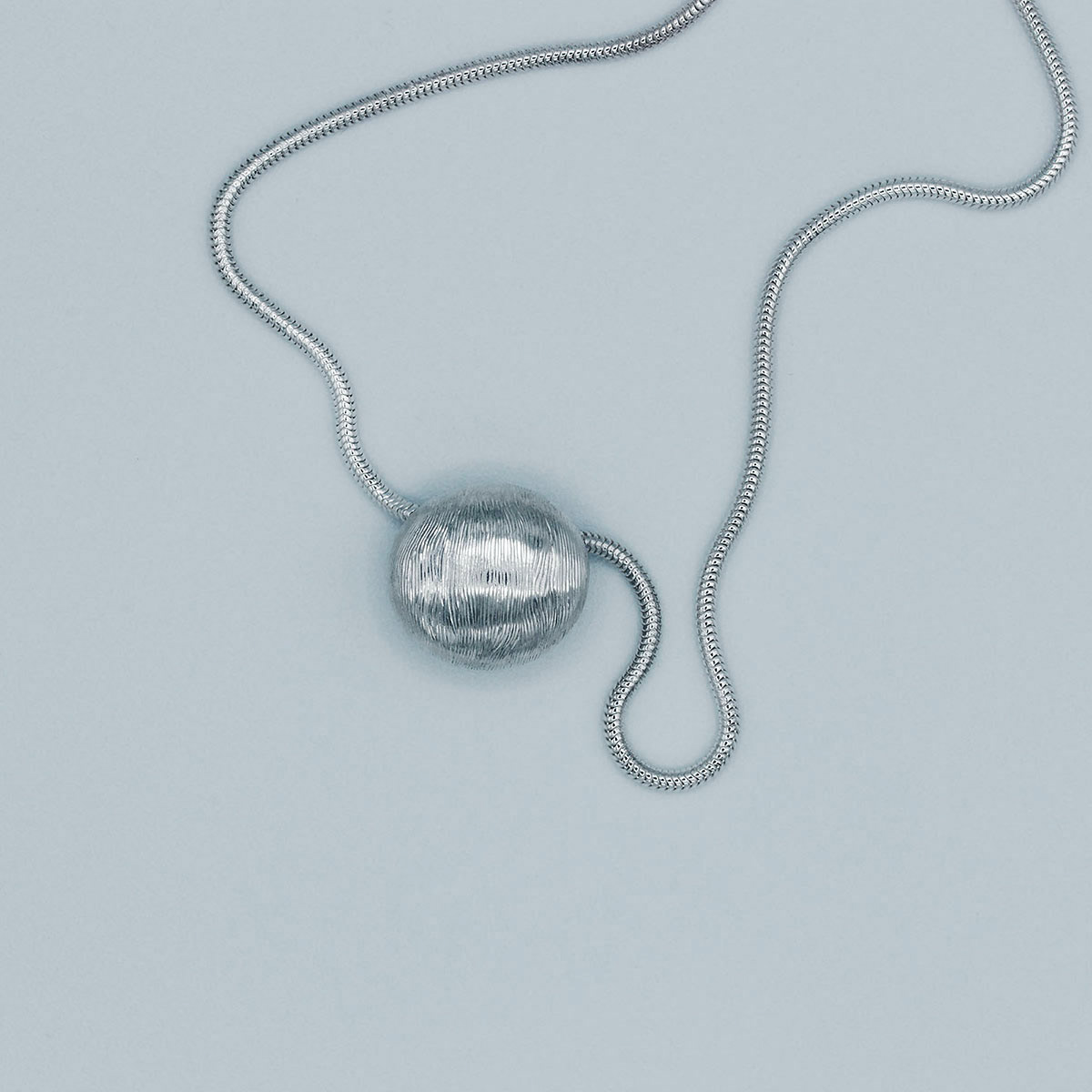 Handcrafted silver pendant with intricate hand-engraved wood-like texture, featuring a unique free-flowing pattern and complemented by a serpentine chain. | Pendentif en argent fait main avec une texture finement gravée rappelant le bois, présentant un motif unique fluide et accompagné d'une chaîne serpentine.