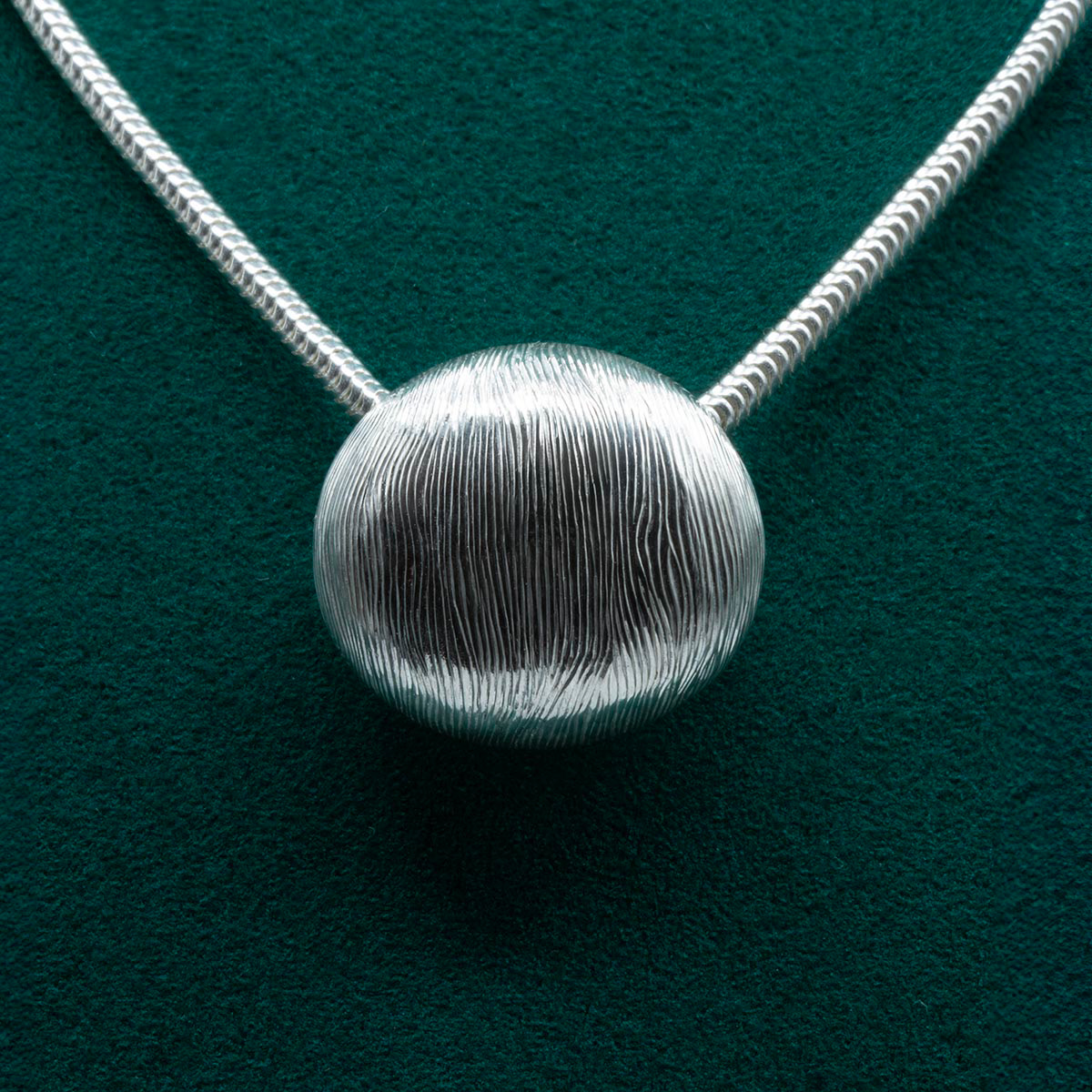 Handcrafted silver pendant with intricate hand-engraved wood-like texture, featuring a unique free-flowing pattern and complemented by a serpentine chain. Pendentif en argent fait main avec une texture finement gravée rappelant le bois, présentant un motif unique fluide et accompagné d'une chaîne serpentine.