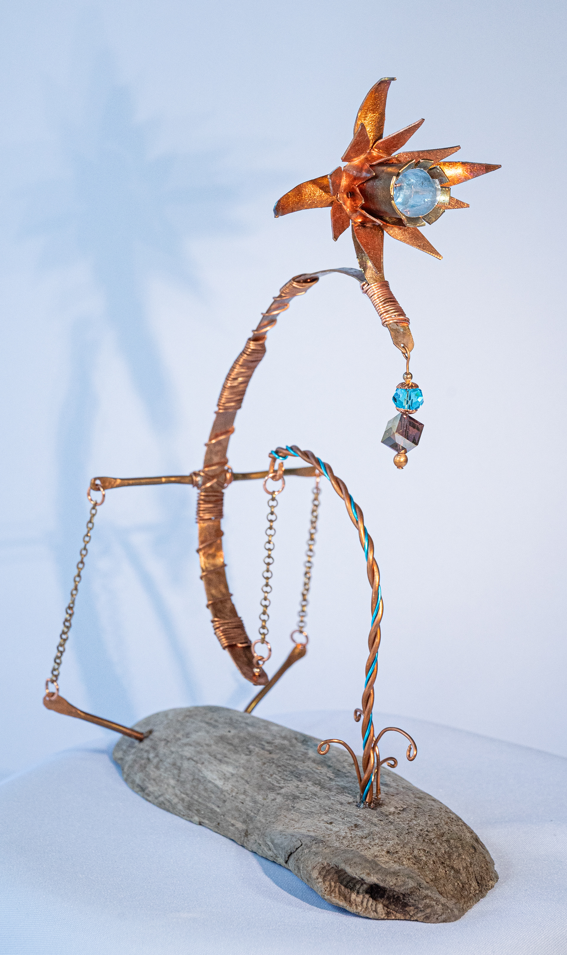 "Weightless" features a gravity held copper and aluminum flower with copper stem, hand twisted wire, brass chain, iridescent and blue crystals on a driftwood base, 10 inches high x 8 inches wide x 4 inches deep, $175