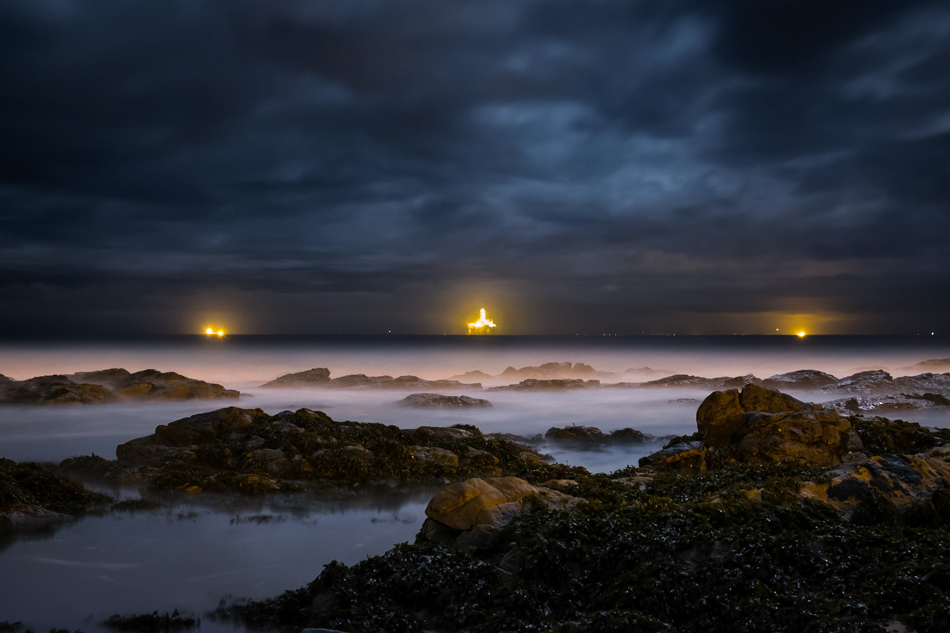 An oil platform and the lights of the Moray coast provide a backdrop to the moonlit Moray Firth at Balintore.