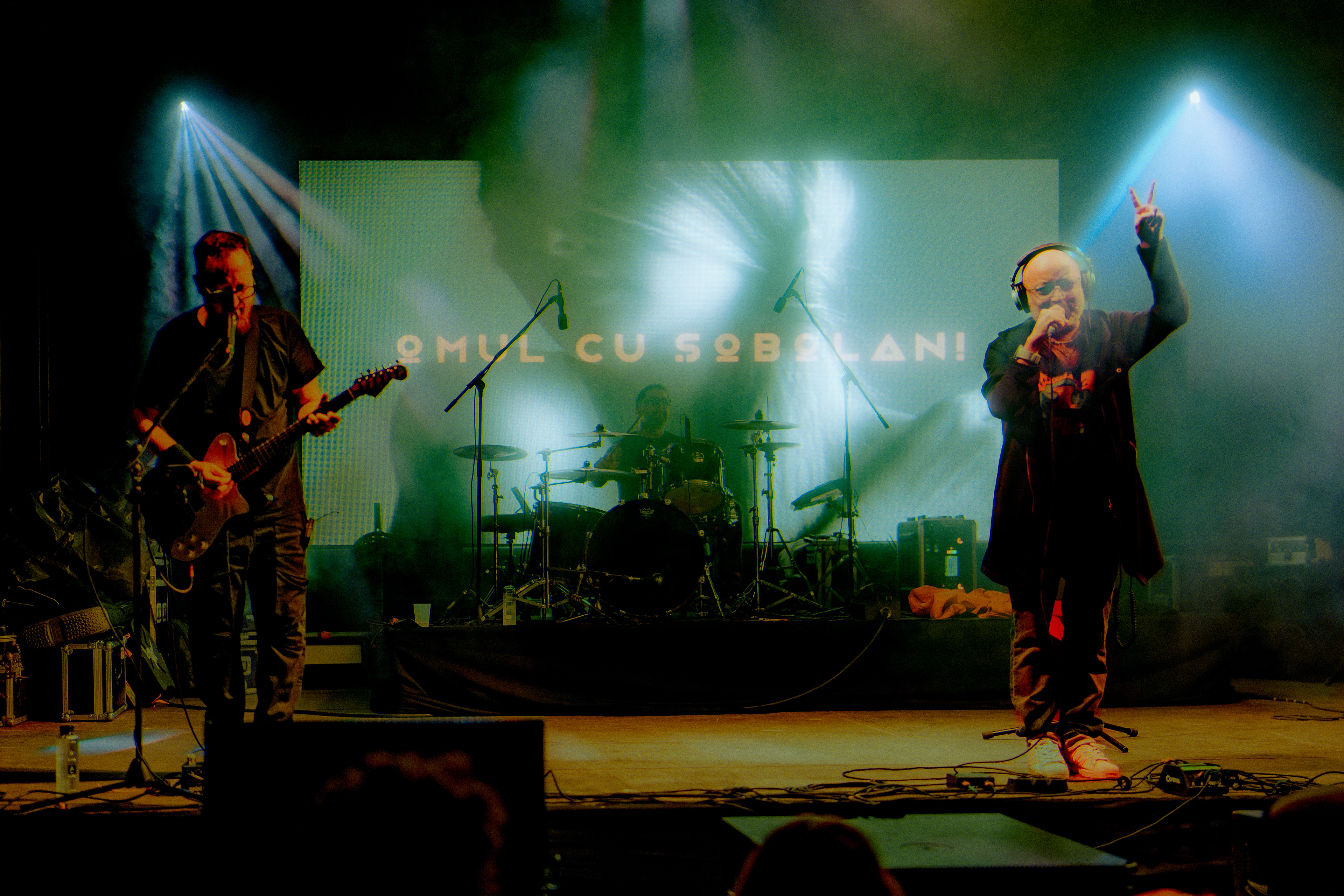 Omul cu Șobolani performing "Două Beri Goale," vocalist holding two fingers in the air, captured in dynamic live performance photography.
