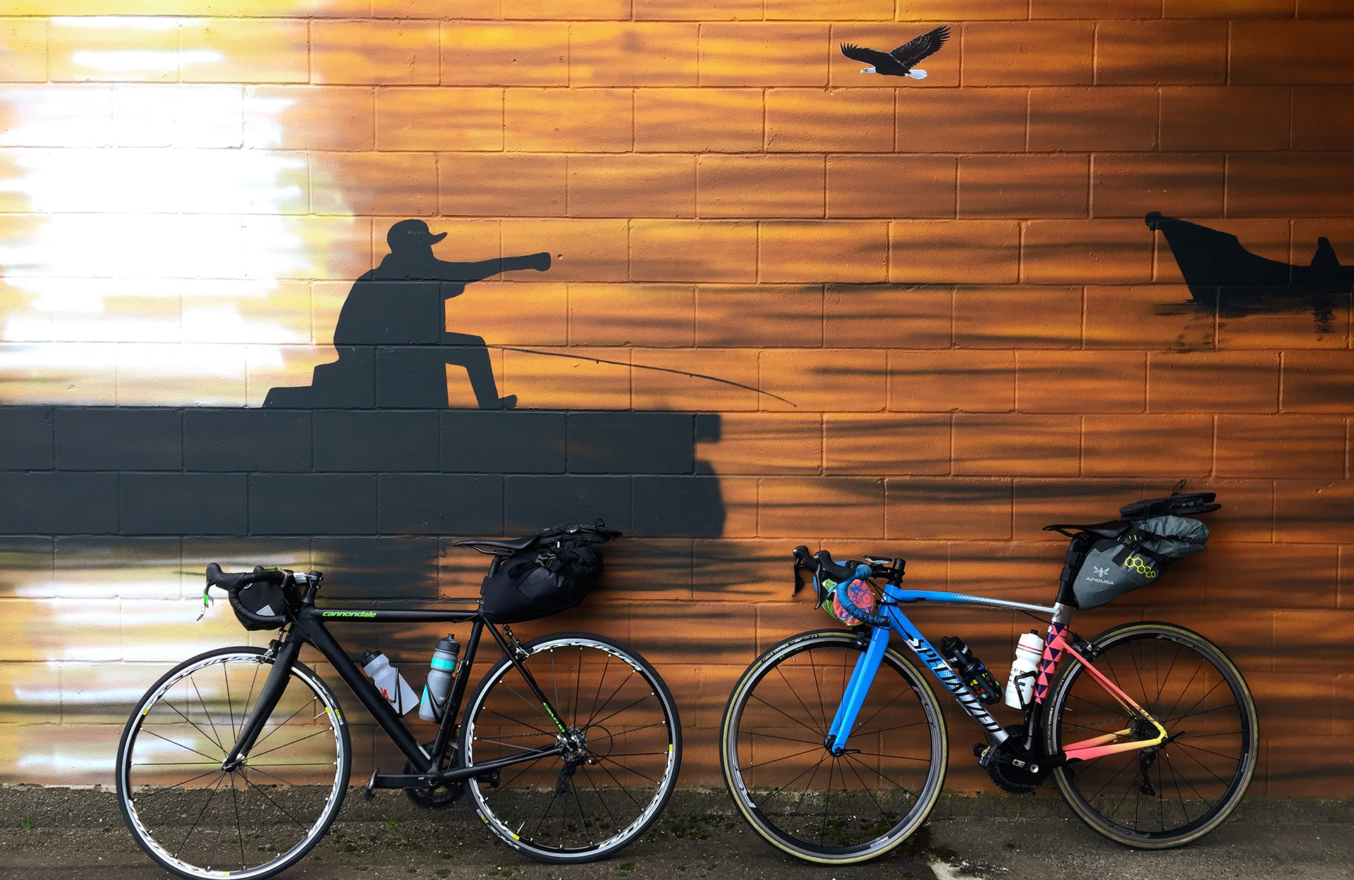 Two road bicycles leaning up against a wall mural of a person fishing.