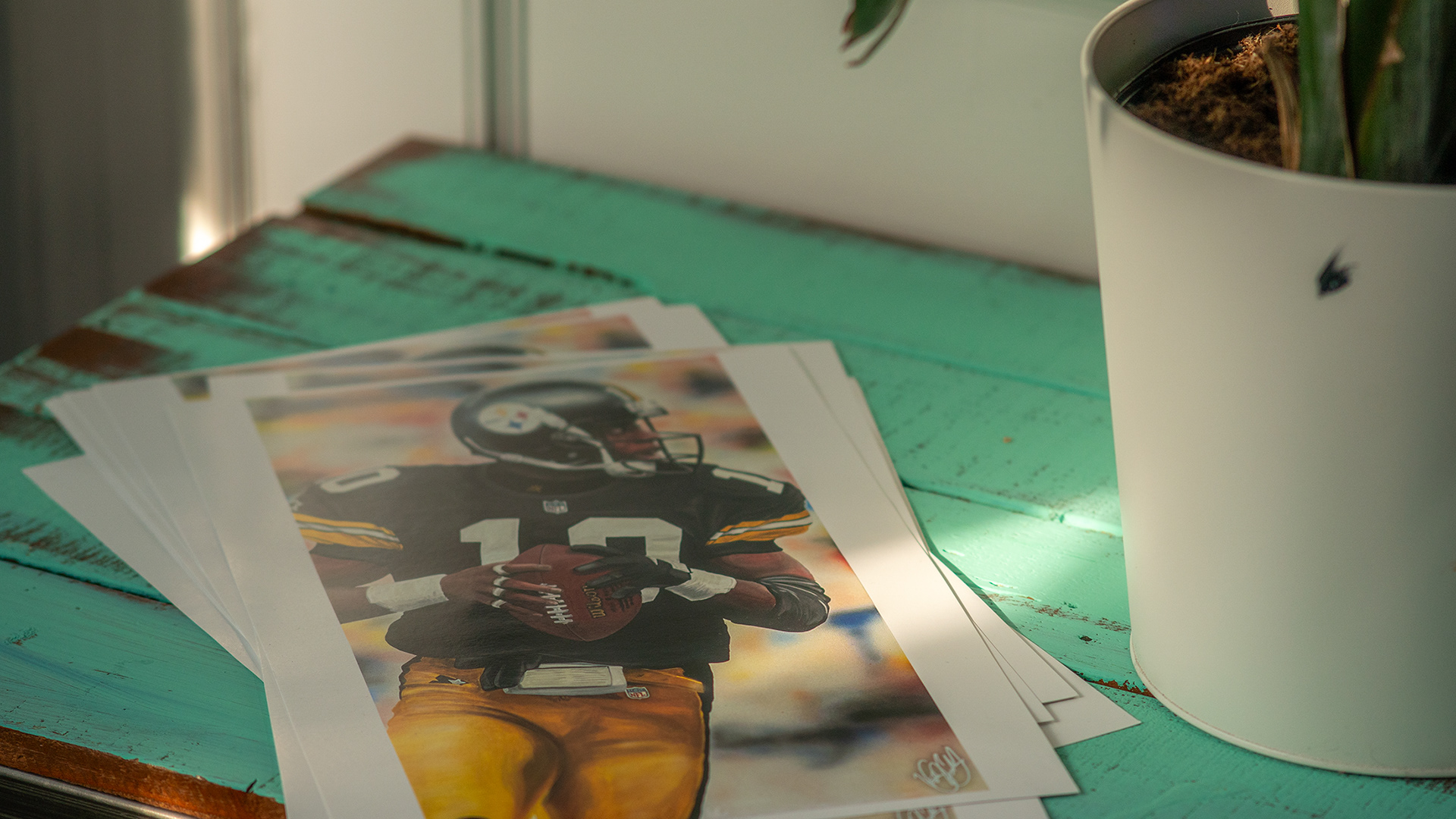 Printed photo of a painting of Kordell Stewart, sitting on a seafoam distressed table, plant in a plain vase on the right hand side, sun shining on the table through a window not pictured