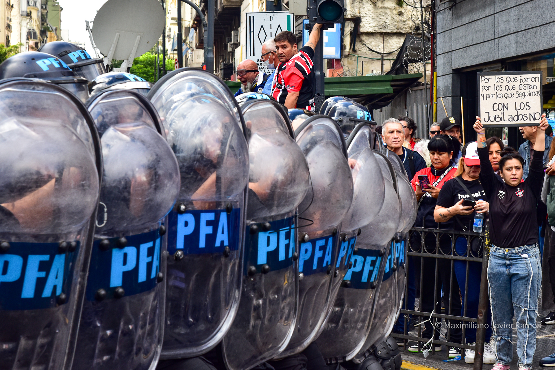 A police formation faces the protesters. On a corner, a woman holds a sign that reads: "For those who were, for those who are, and for those of us who remain. Don't mess with the retirees." In the background, soccer fans who supported the retirees during the march. // Una formación policial se enfrenta a los manifestantes. En una esquina, una mujer sostiene un cartel que dice: "Por los que fueron, por los que están y por los que quedamos. Con los jubilados no". Al fondo, aficionados al fútbol que apoyaron a los jubilados durante la marcha.