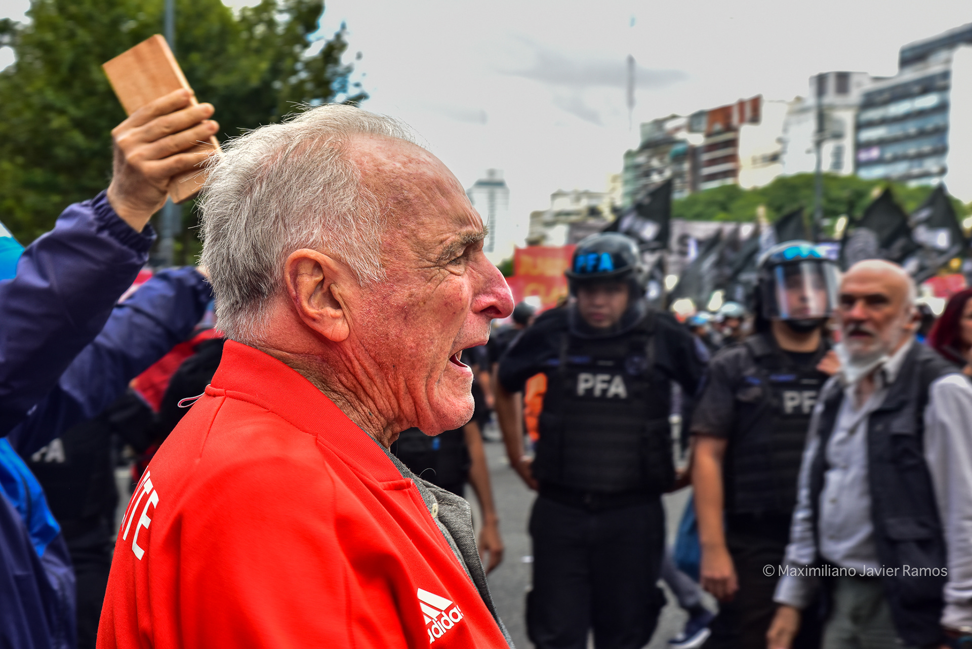 Un jubilado grita cerca del Congreso Nacional Argentino durante una manifestación conjunta entre jubilados e hinchas de fútbol. Al fondo se ve a policías intentando despejar la zona. // A retiree shouts near the Argentine National Congress during a joint demonstration between retirees and soccer fans. Police officers are seen in the background trying to clear the area.