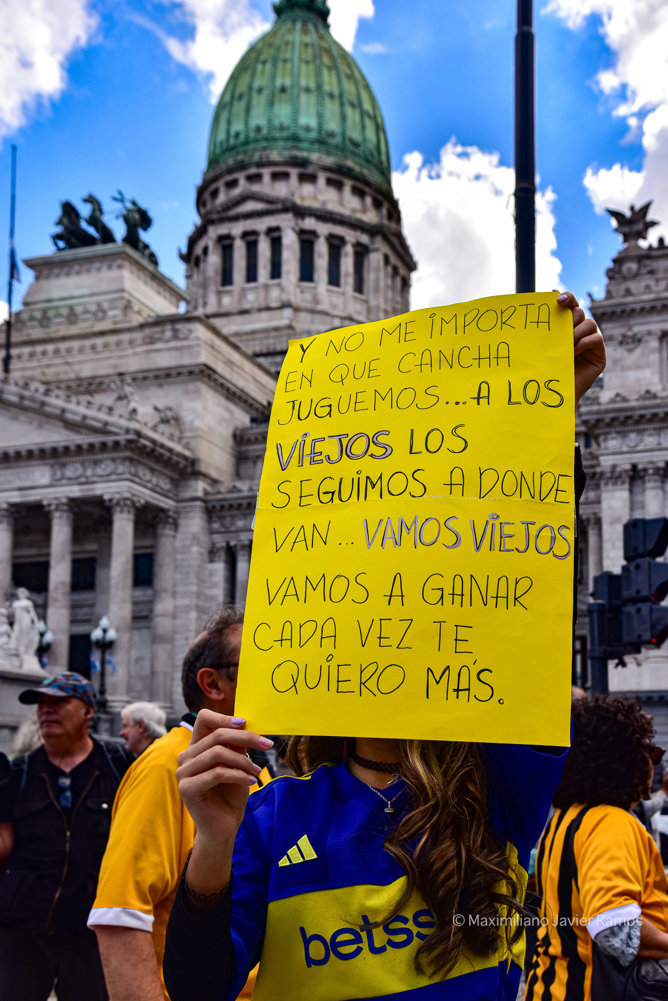 Una joven hincha de Boca Juniors sostiene un cartel que dice: "Y no me importa en qué estadio juguemos... seguimos a los viejos adonde vayan... ¡Vamos, viejos, vamos a ganar! Los quiero cada vez más". Al fondo, el Congreso Nacional Argentino. // A young Boca Juniors fan holds a sign that reads, "And I don't care what stadium we play on... we follow the old guys wherever they go... come on, old guys, we're going to win. I love you more and more." The Argentine National Congress is in the background.