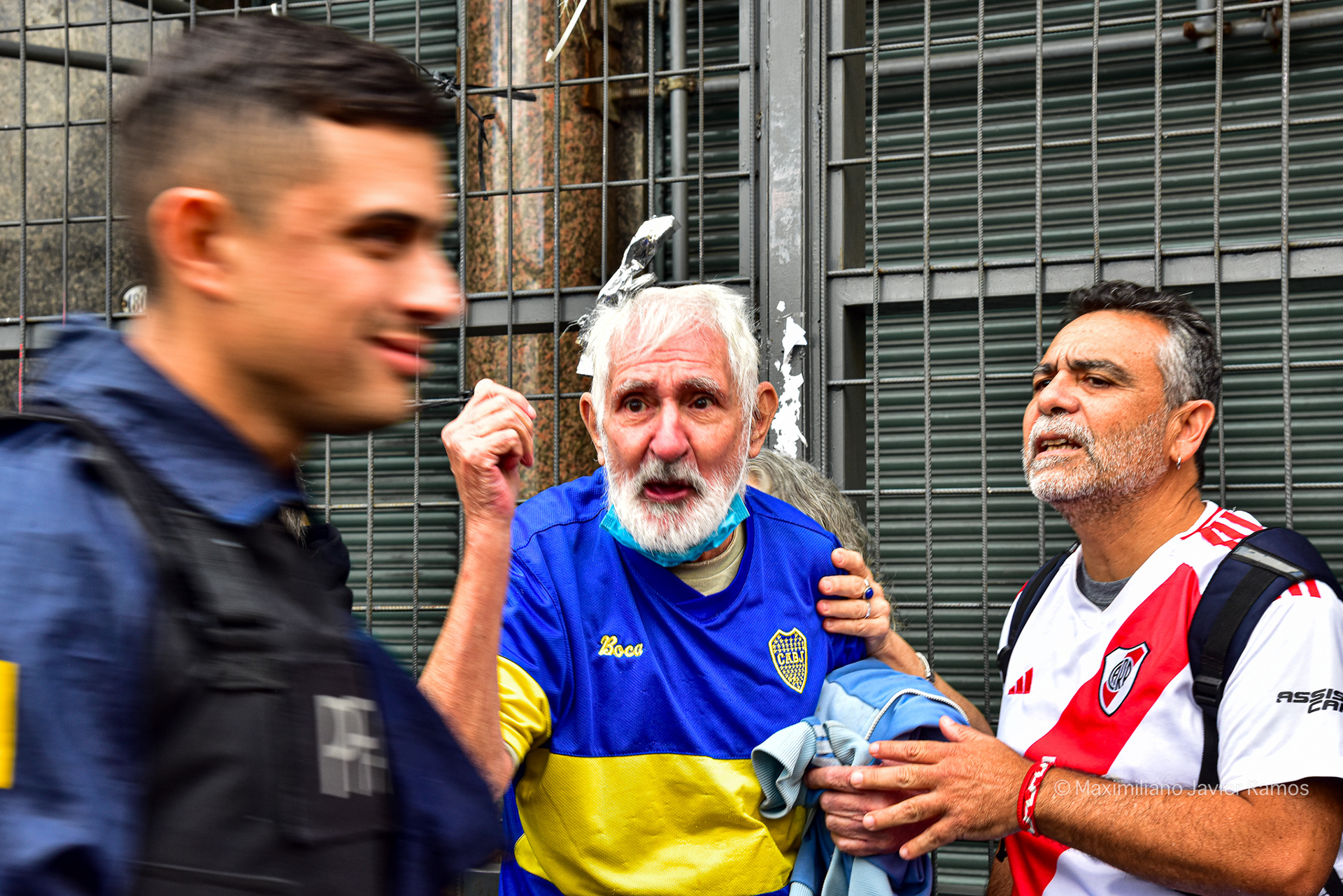 Dos jubilados, con camisetas de Boca y River Plate, le gritan a un policía que pasa riendo mientras, a pocos pasos, la infantería policial reprime a los manifestantes. // Two retirees, wearing Boca Juniors and River Plate jerseys, shout at a passing police officer, laughing, while, a few steps away, police troops repress the protesters.