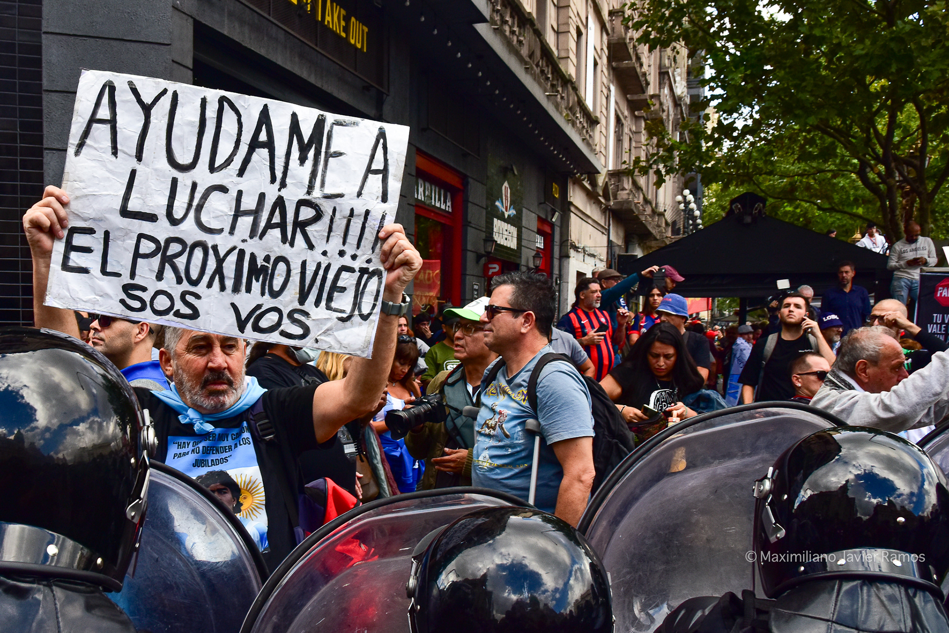 Un jubilado sostiene, con un cordón de infantería de la Gendarmería Nacional al fondo, dos carteles que dicen: "Nos robaron todo a los viejos, menos la moral de la lucha. ¡Vengan, únanse a los jubilados, hay que echar a la mafia! ¡Maldita sea!" y otro: "Policía corrupta, violenta y asesina". - Un Jubilado sostiene, frente a la policía, un cartel que dice: "¡Ayúdame a luchar! ¡El próximo viejo sos vos!". //