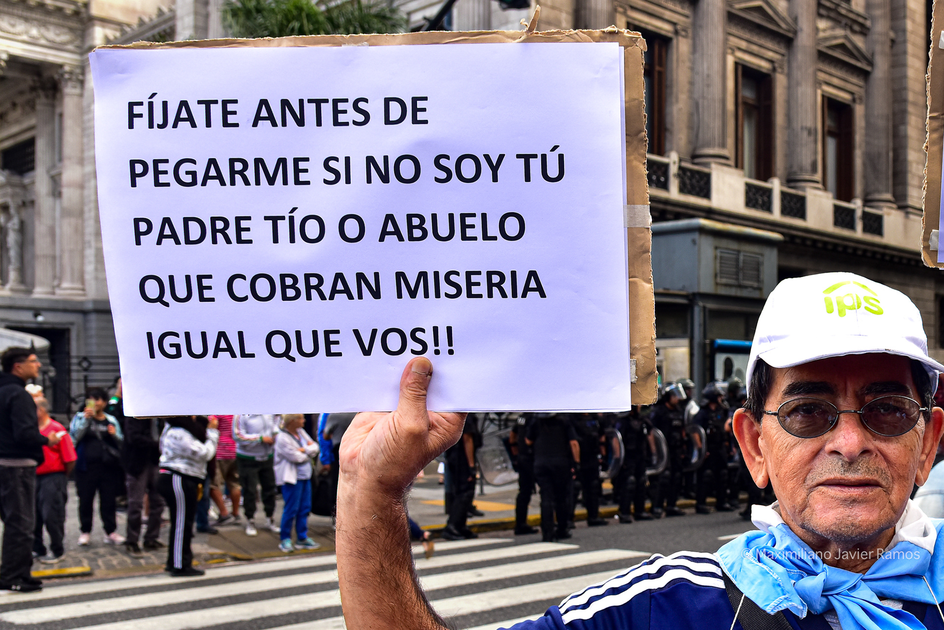 Frente al Congreso Nacional, con la policía al fondo, un jubilado sostiene un cartel que dice: "Antes de pegarme, fíjate si no soy tu padre, tío o abuelo, que gana una miseria igual que vos". // In front of the National Congress, with the police in the background, a retiree holds a sign reading: "Before hitting me, check if I'm not your father, uncle, or grandfather who earns a pittance just like you".