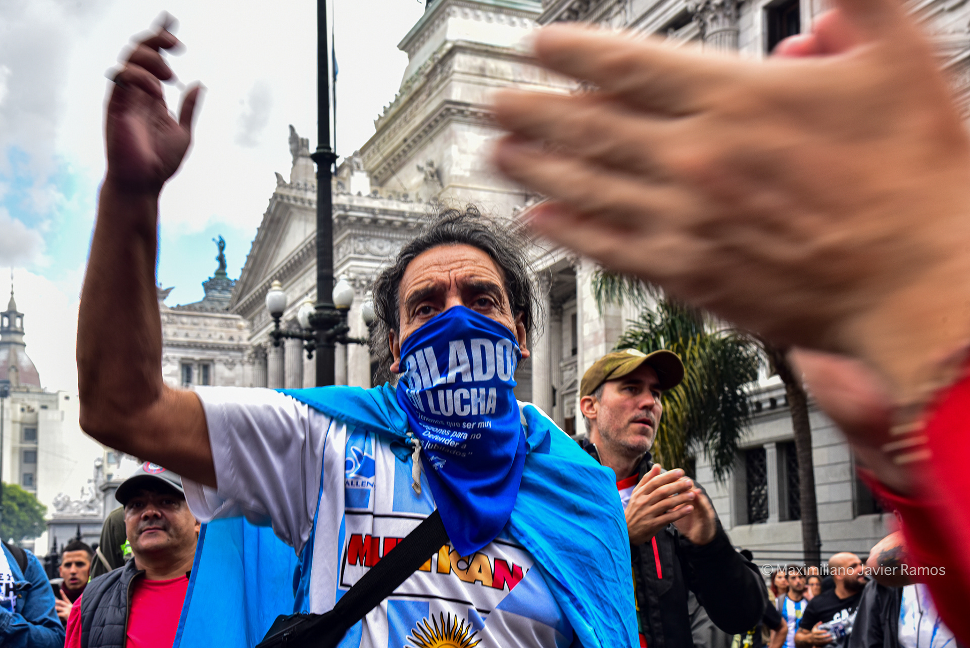 Un hombre con el rostro cubierto por una bufanda que dice "Jubilados en Lucha" se manifiesta con el Congreso Nacional al fondo. // A man with his face covered by a scarf that says "Retirees in Struggle" demonstrating with the National Congress in the background.