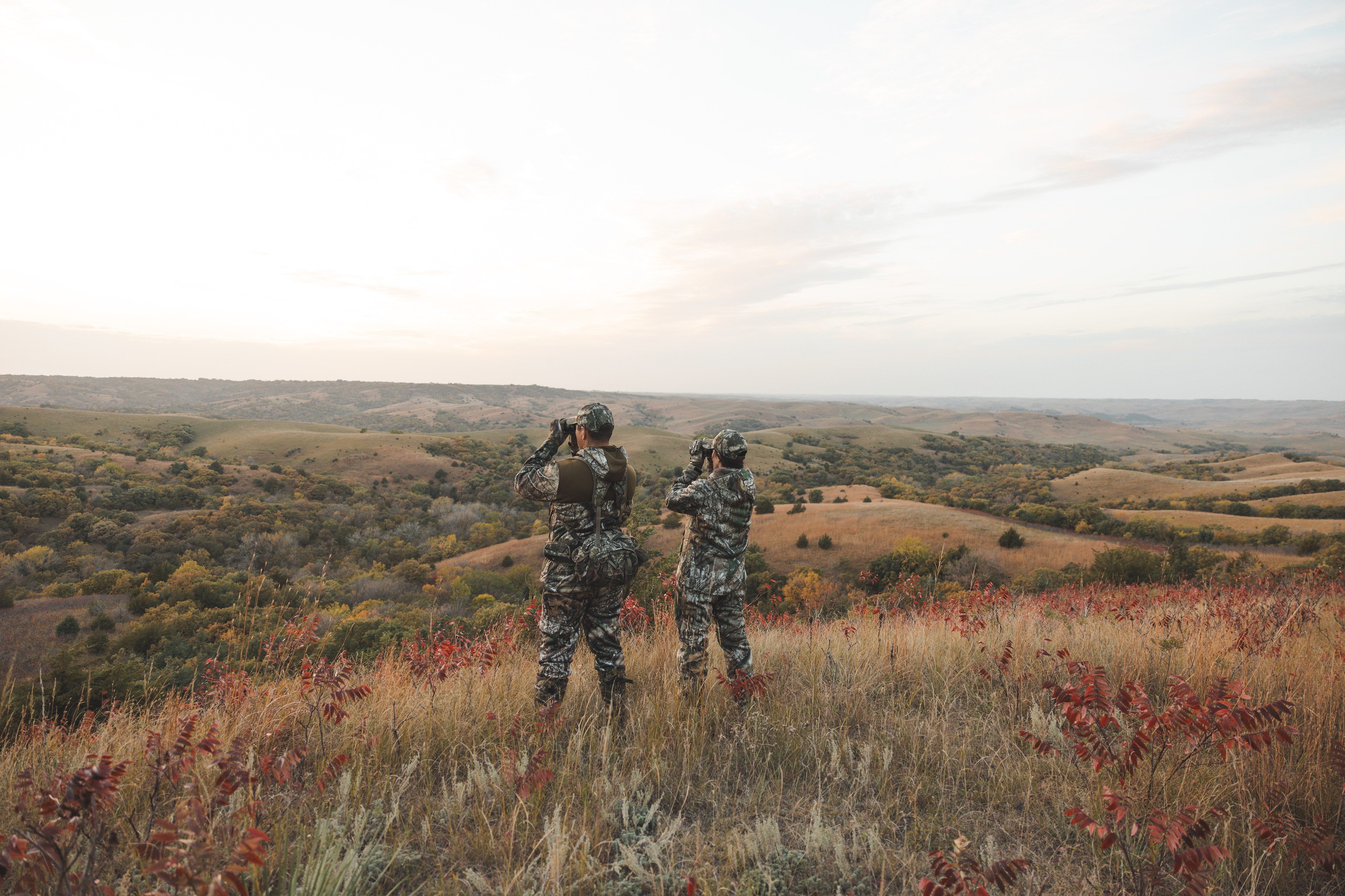 Two hunters dressed in Realtree camoflage standing on a hill, looking over the South Dakota landscape through binoculars 