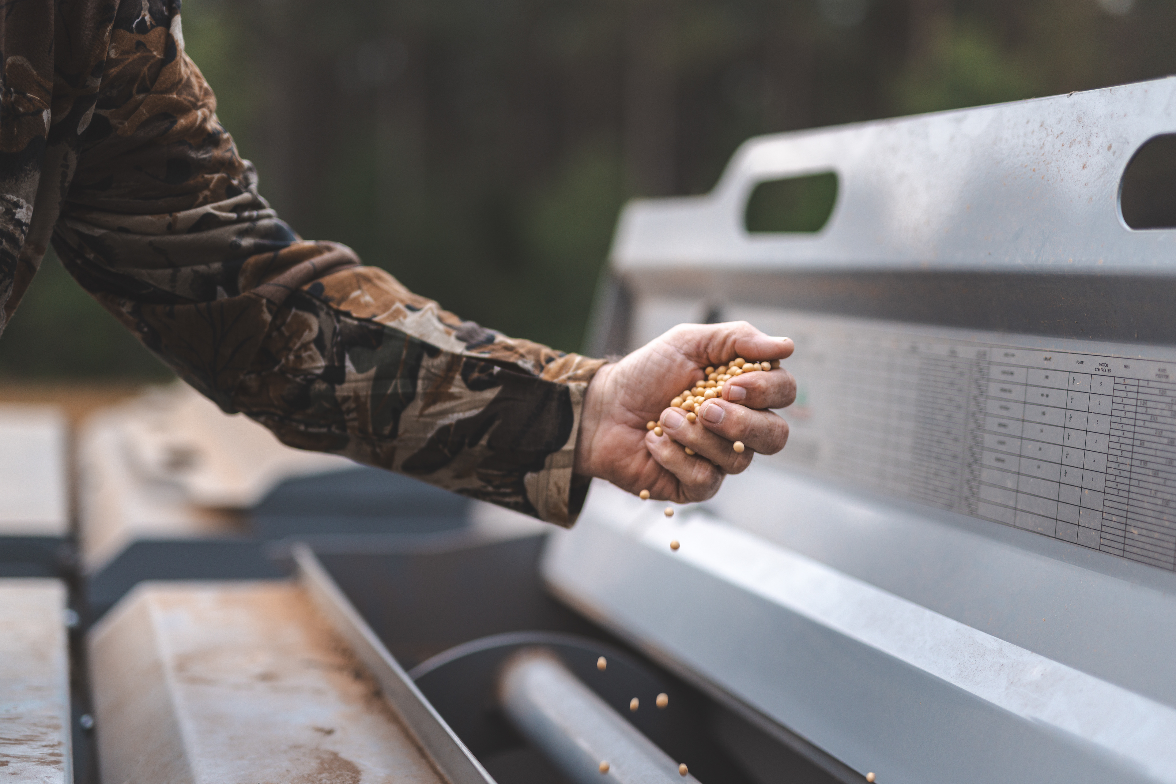 an arm stretched out over a seeder of a tractor, dropping in clover seeds while wearing a Realtree camo shirt