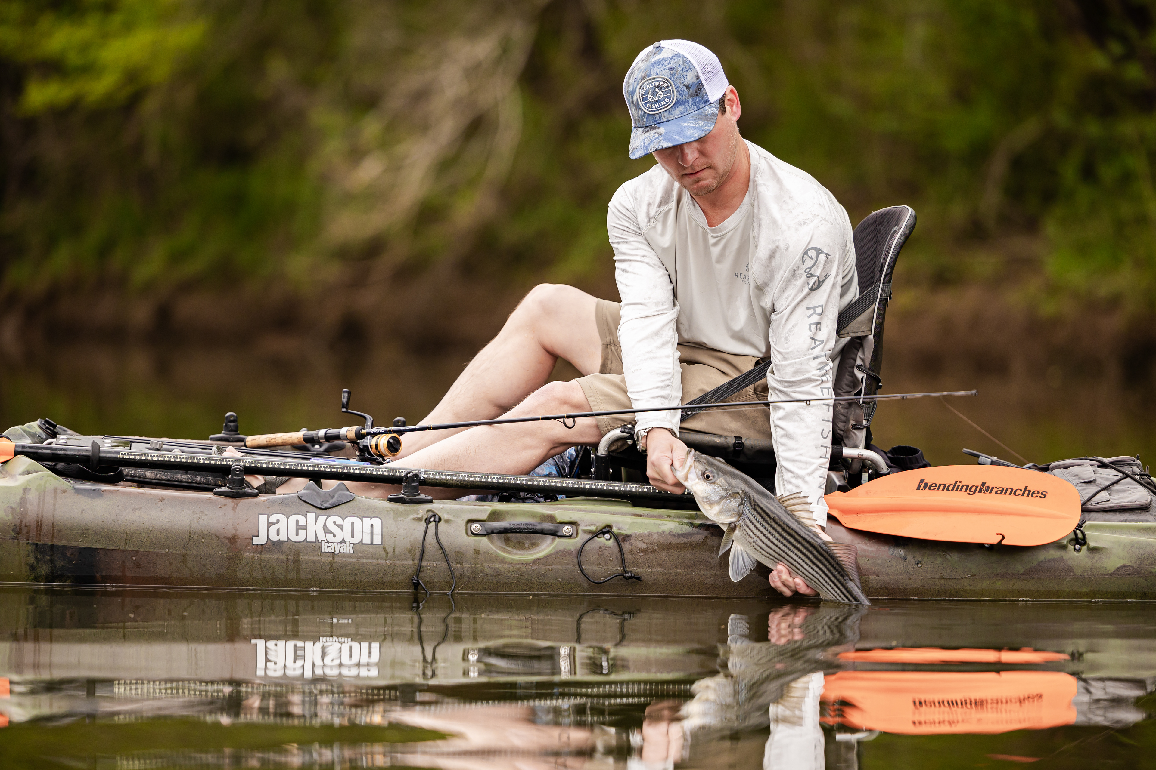 Fly Fishing Angler wearing a Realtree fishing shirt and Realtree fishing hat on a kayak , pulling up a striped bass. On the Tallapoosa River