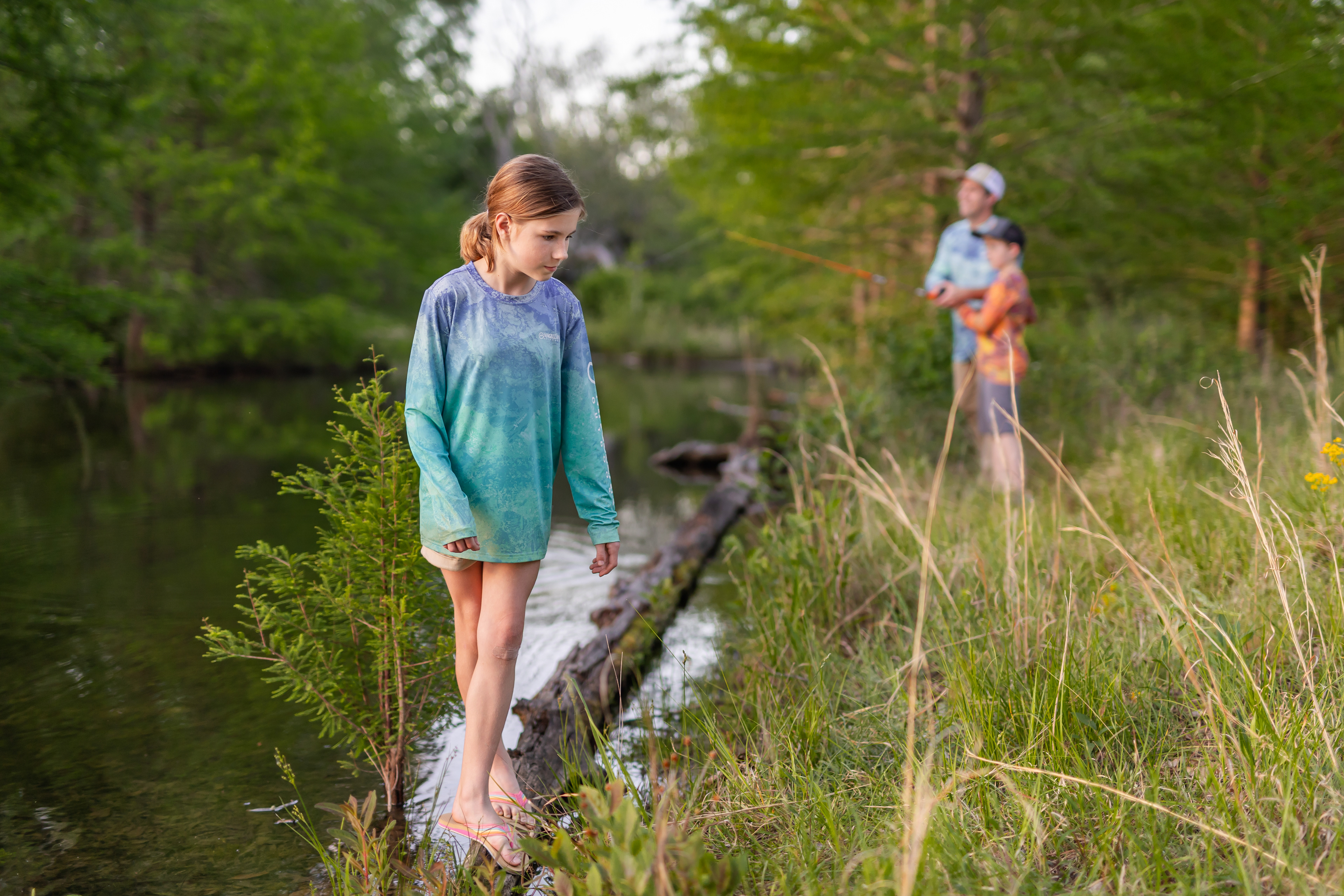 Girl in a Magellan Realtree shirt balancing on a log, while the father and brother are fishing in the background while wearing Realtree fishing shirts